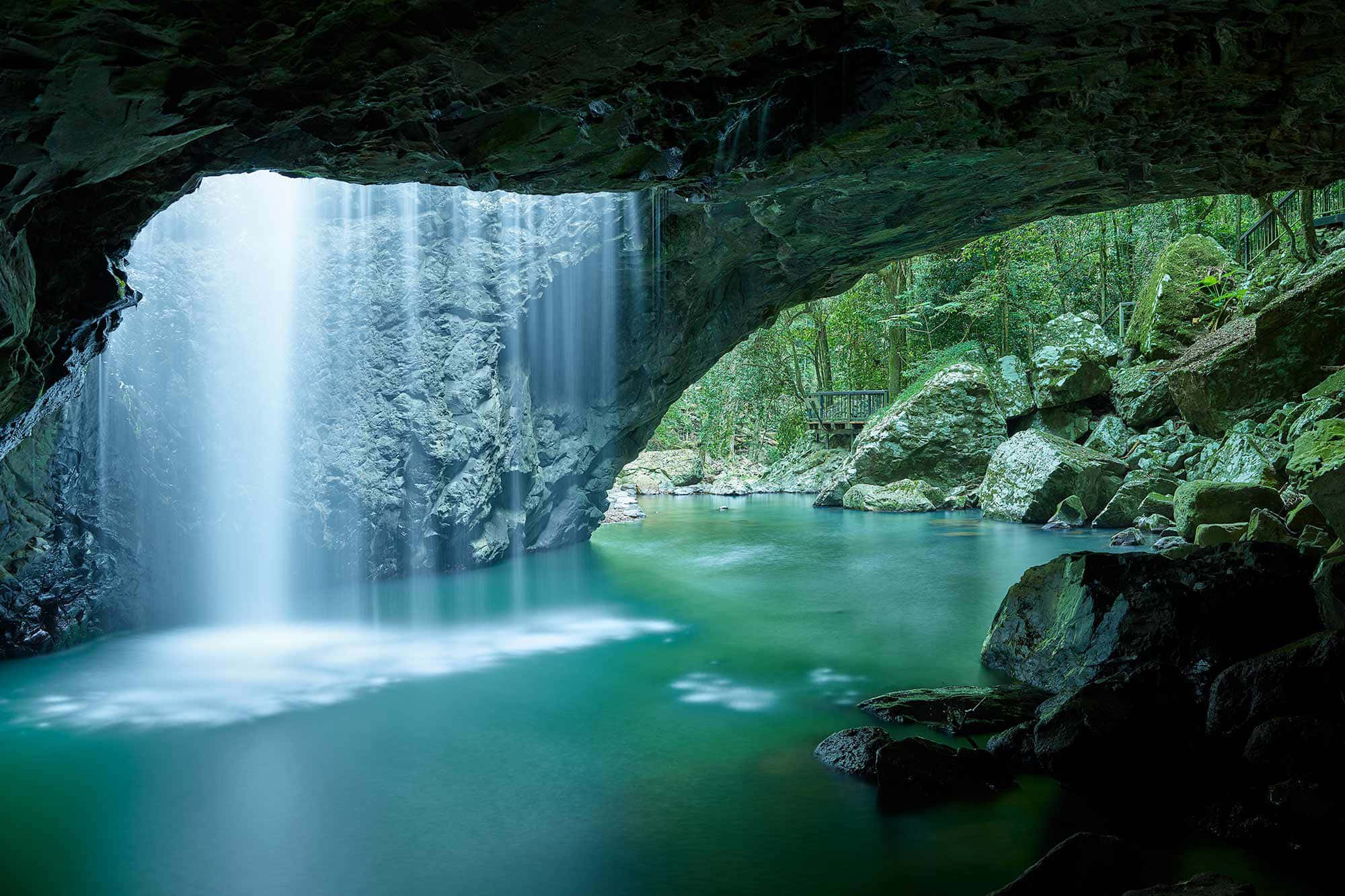 洞穴 桌布 壁紙 — 一道寧靜的瀑布穿過天然岩石拱門，流入被茂密綠色森林環繞的碧綠水池。
