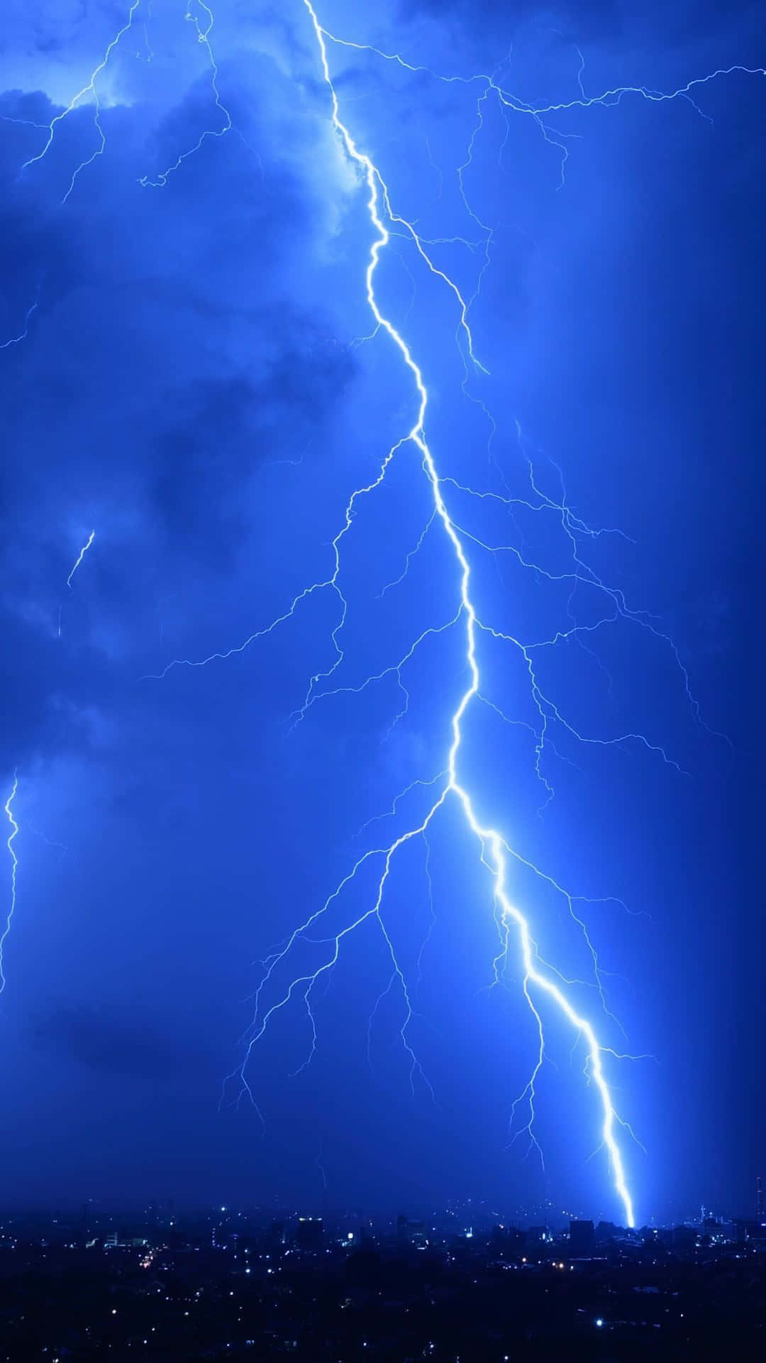 Thunderstorm 桌布 壁紙 — 一道巨大的閃電在深藍色暴風雨天空中分叉，下方是閃爍著遠方燈光的城市景觀。