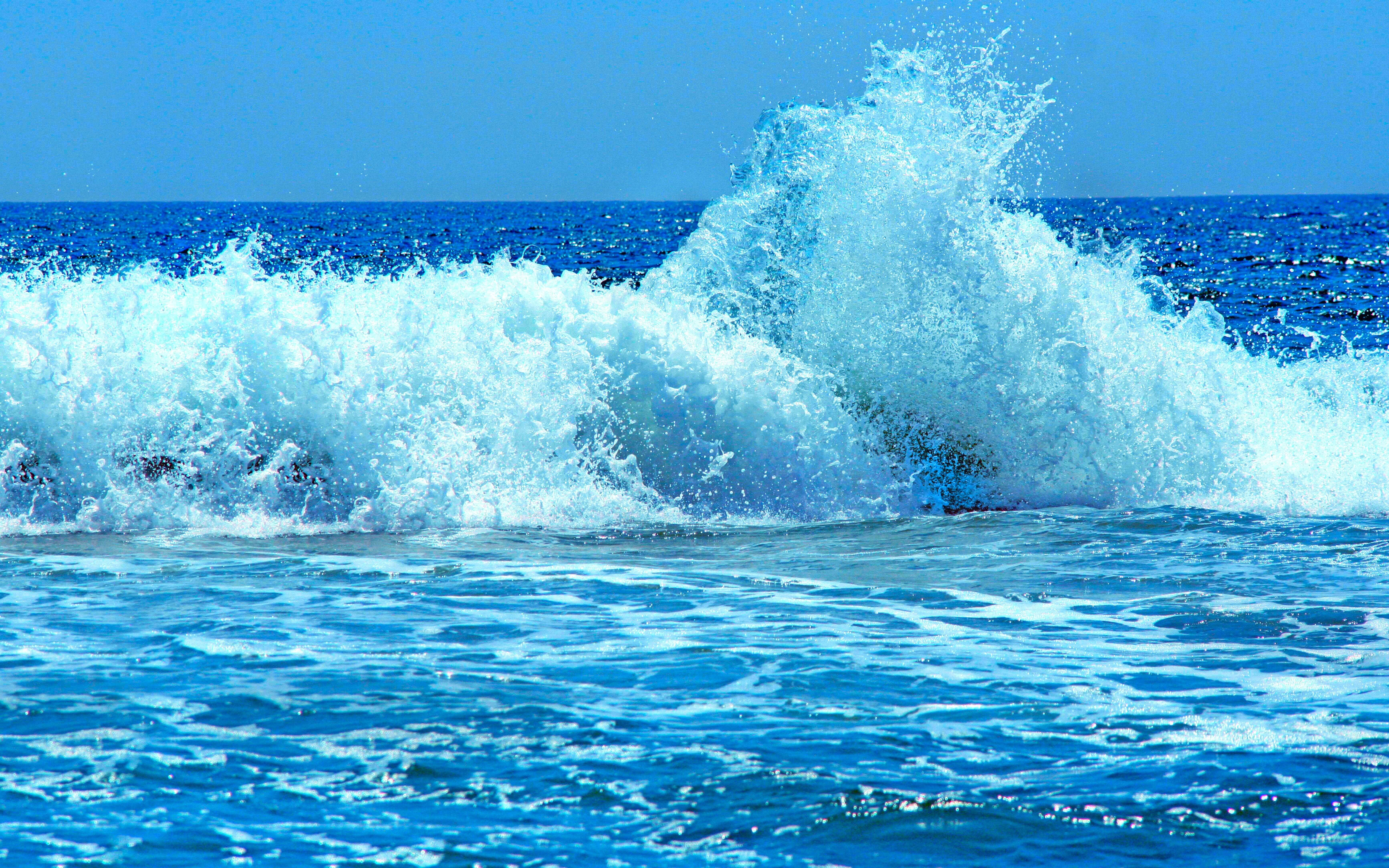 waves 桌布 壁紙 — 一道強勁的海浪帶著白色泡沫拍打岸邊，天空清澈湛藍。