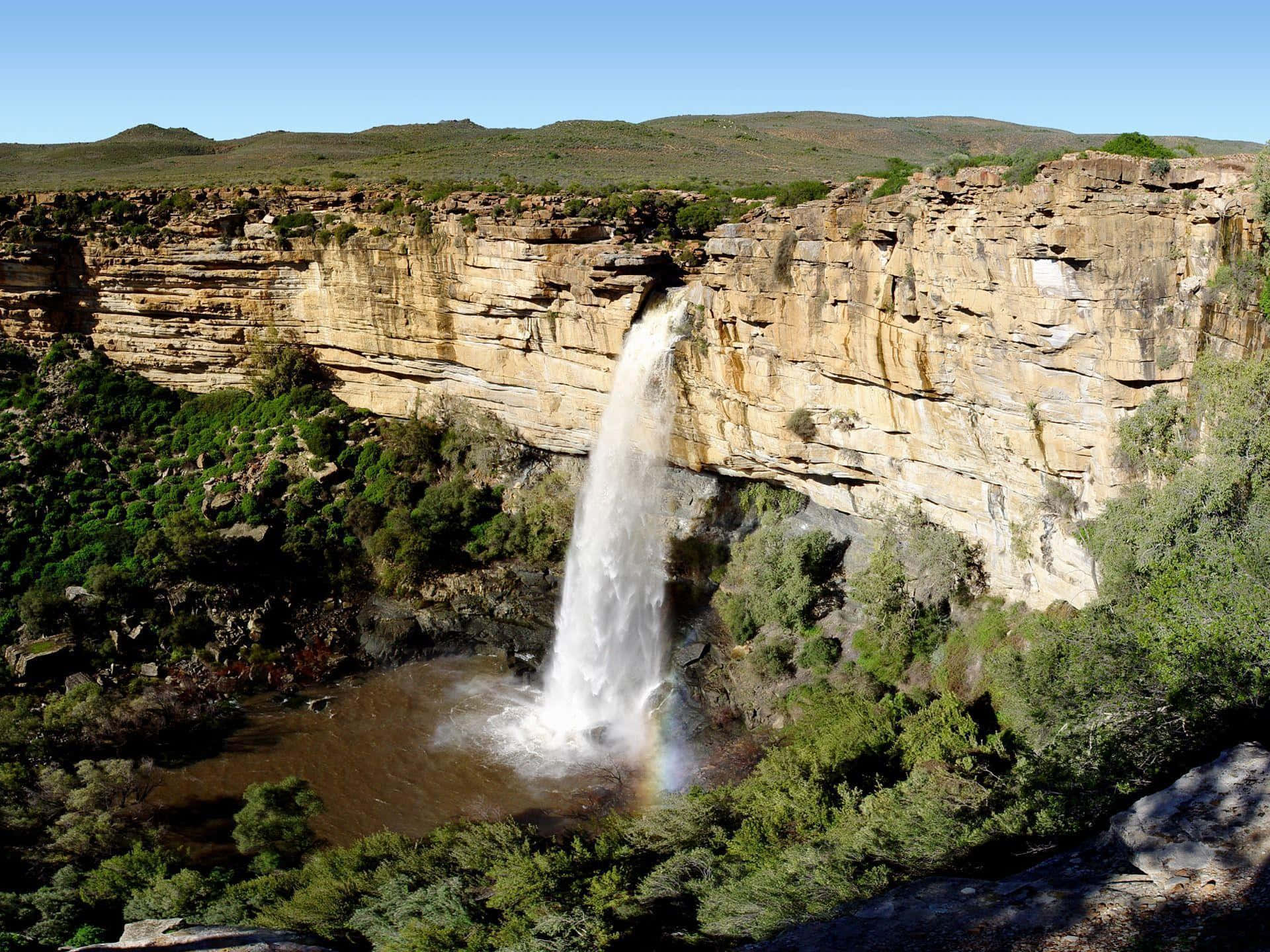 South Africa 桌布 壁紙 — 一道強勁的瀑布從崎嶇的懸崖傾瀉而下，落入煙霧繚繞的水潭，四周環繞著茂密的綠植，上方是清澈的藍天。