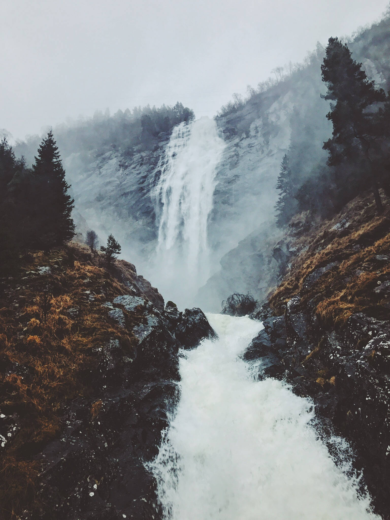 Norway 桌布 壁紙 — 一道強勁的瀑布從霧氣繚繞的懸崖邊傾瀉而下，周圍環繞著深色常青樹和崎嶇的地形。