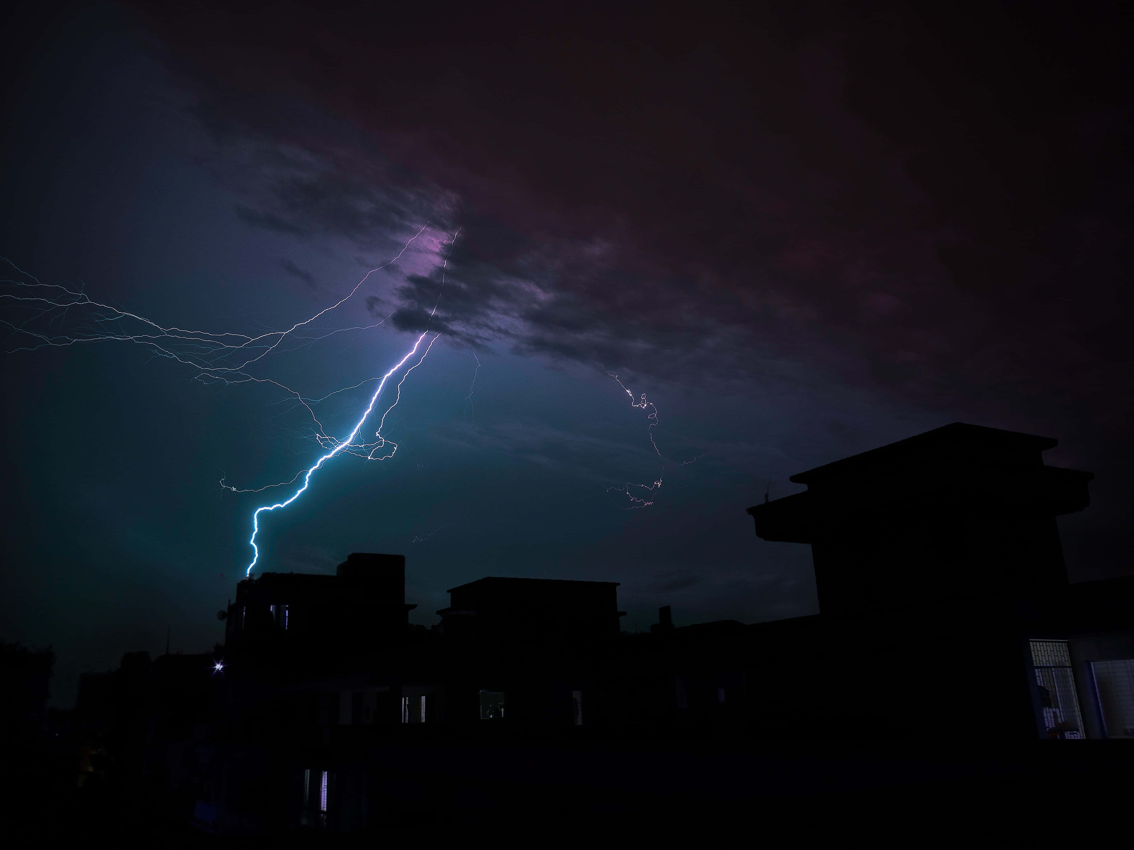 Thunderstorm 桌布 壁紙 — 一道明亮的藍色閃電在夜晚剪影般的城市屋頂上方，於深紫色暴風雨天空中分叉。