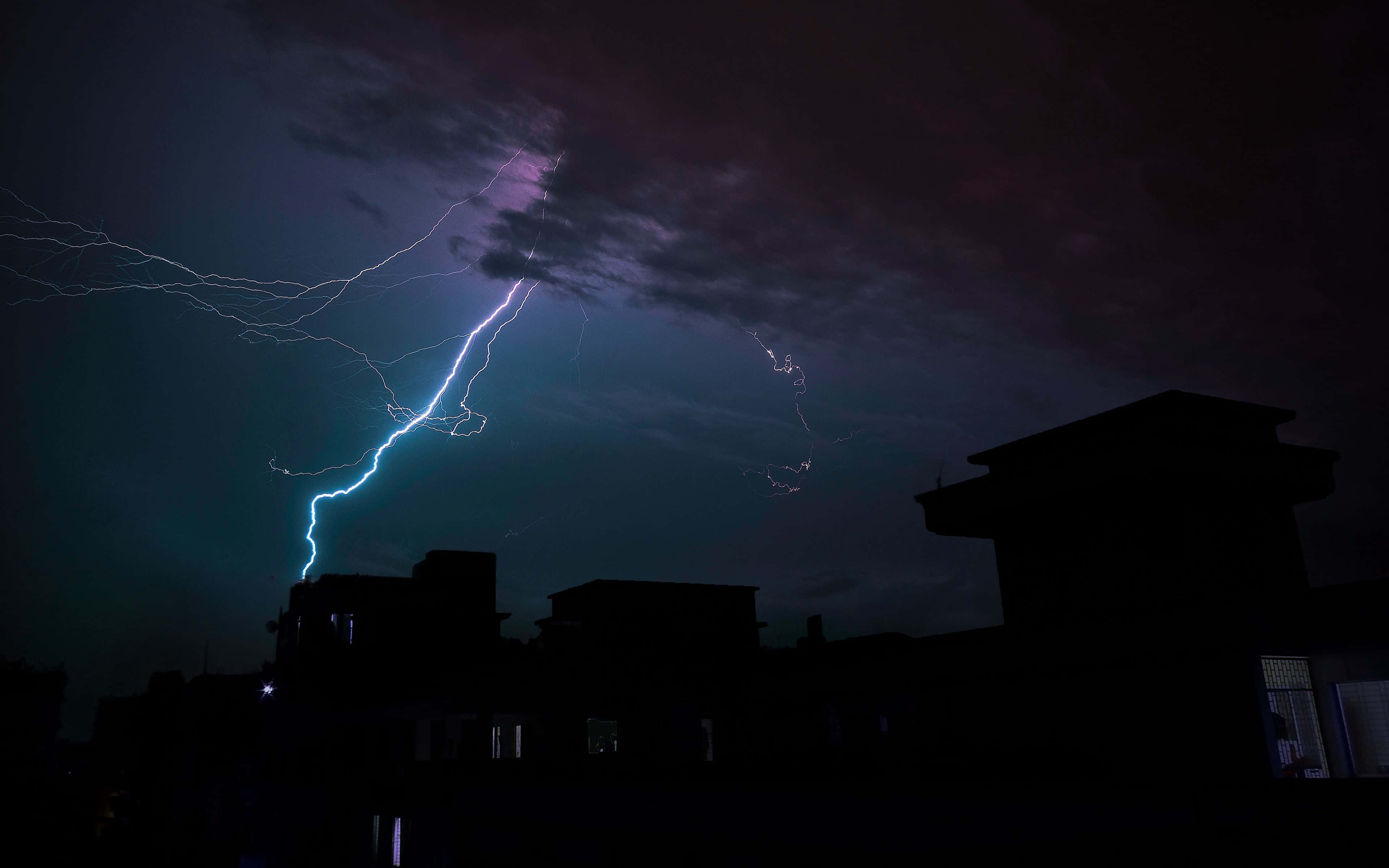 Thunderstorm 桌布 壁紙 — 一道明亮的藍色閃電在夜間剪影般的城市屋頂上方，劃過深紫色的暴風雨天空。