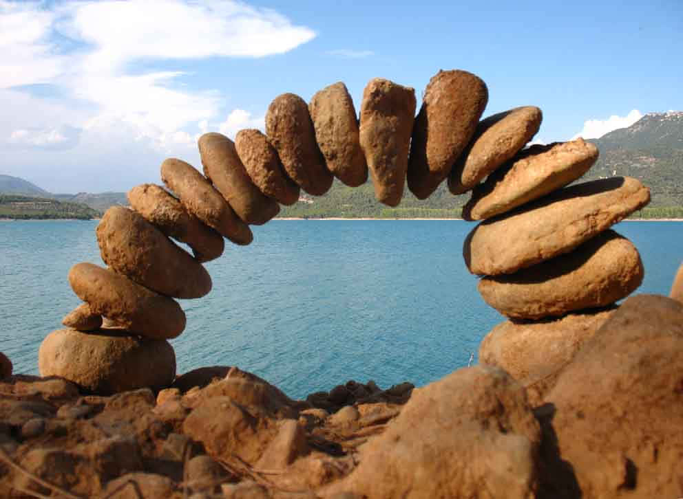 Land Art 桌布 壁紙 — 一道由平衡的棕色石頭組成的精緻拱門，框住了在晴朗藍天和散落雲朵下寧靜的綠松石色湖泊。