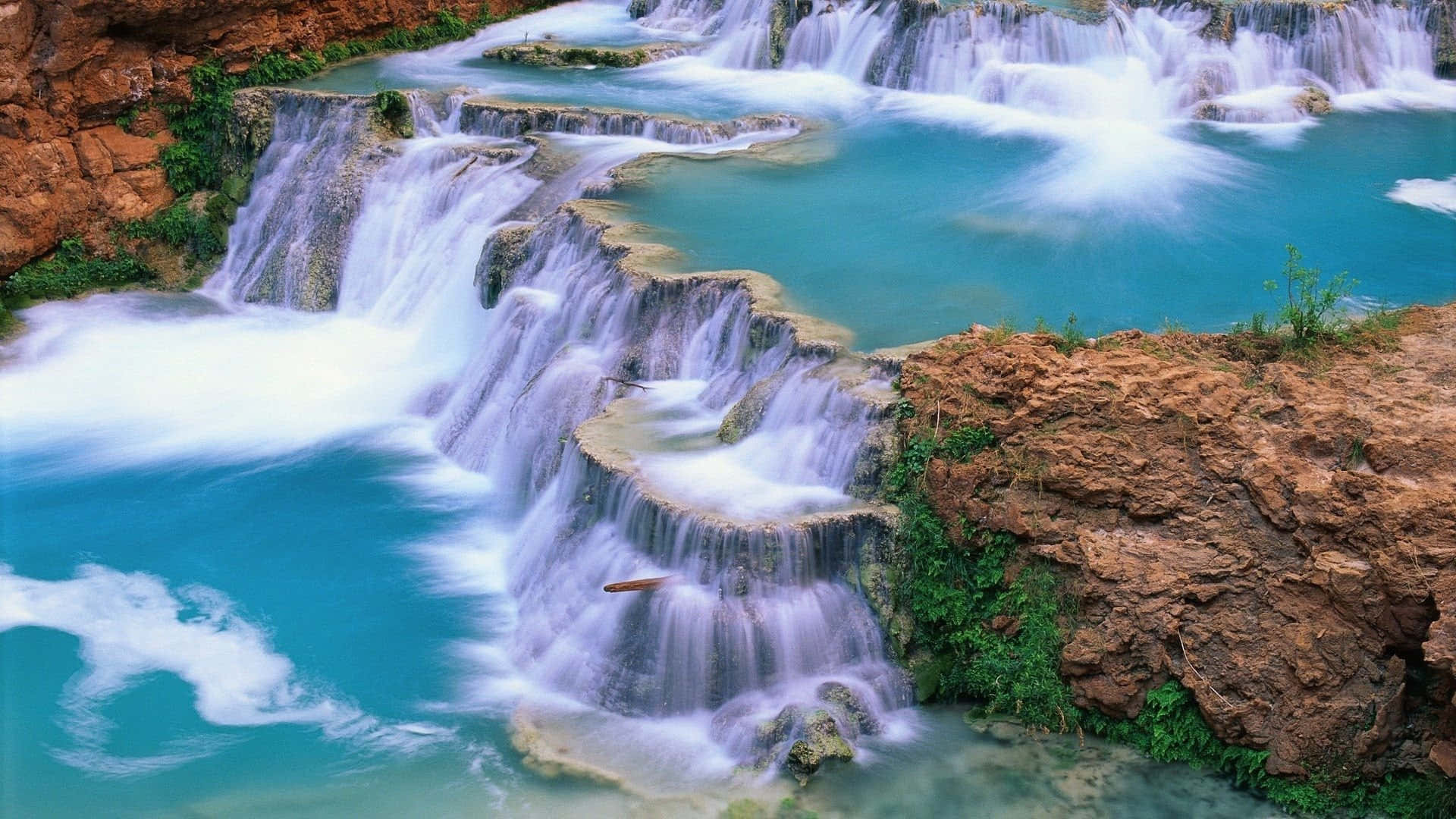 3d Waterfall 桌布 壁紙 — 一道藍綠色瀑布層層疊疊地流過岩石構造，陡峭的懸崖上長滿了茂密的綠色植物。