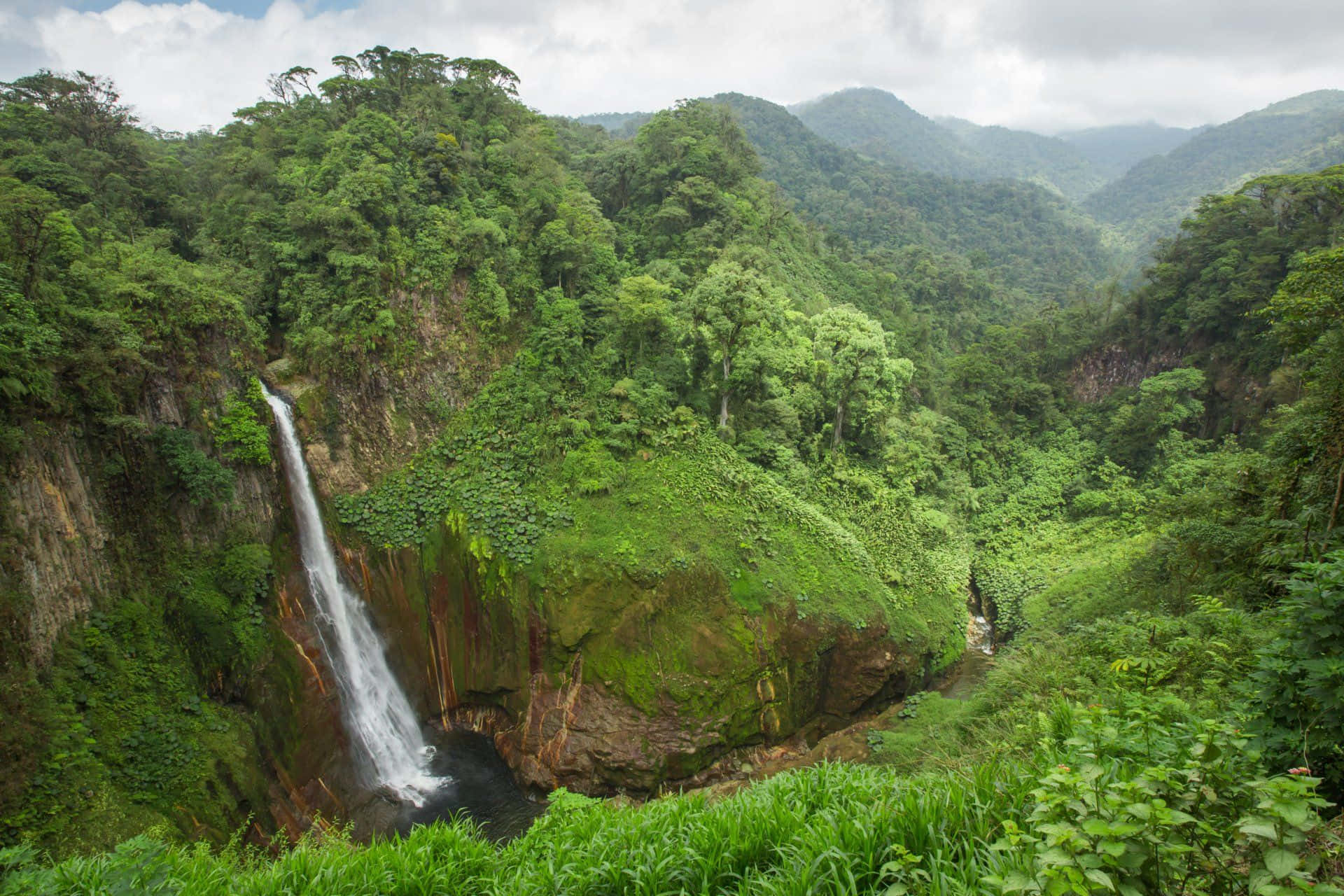 Costa Rica 桌布 壁紙 — 一道雄偉的瀑布從長滿苔蘚的懸崖傾瀉而下，落入一個被茂密翡翠叢林和連綿山脈環繞的水潭，天空多雲。
