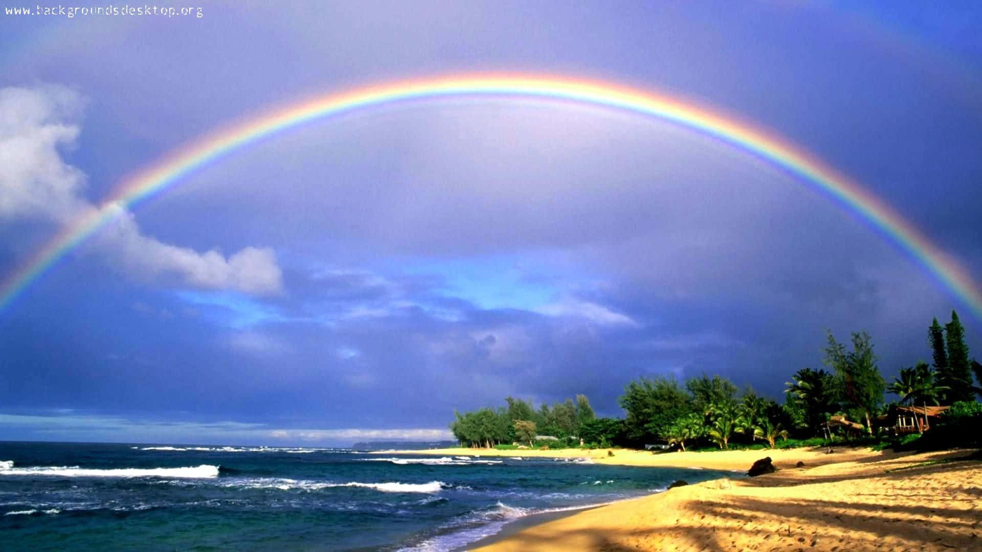 Hawaiian Beach 桌布 壁紙 — 一道鮮豔的彩虹橫跨熱帶海灘，在戲劇性的多雲天空下，碧綠的海浪拍打著金色沙灘。