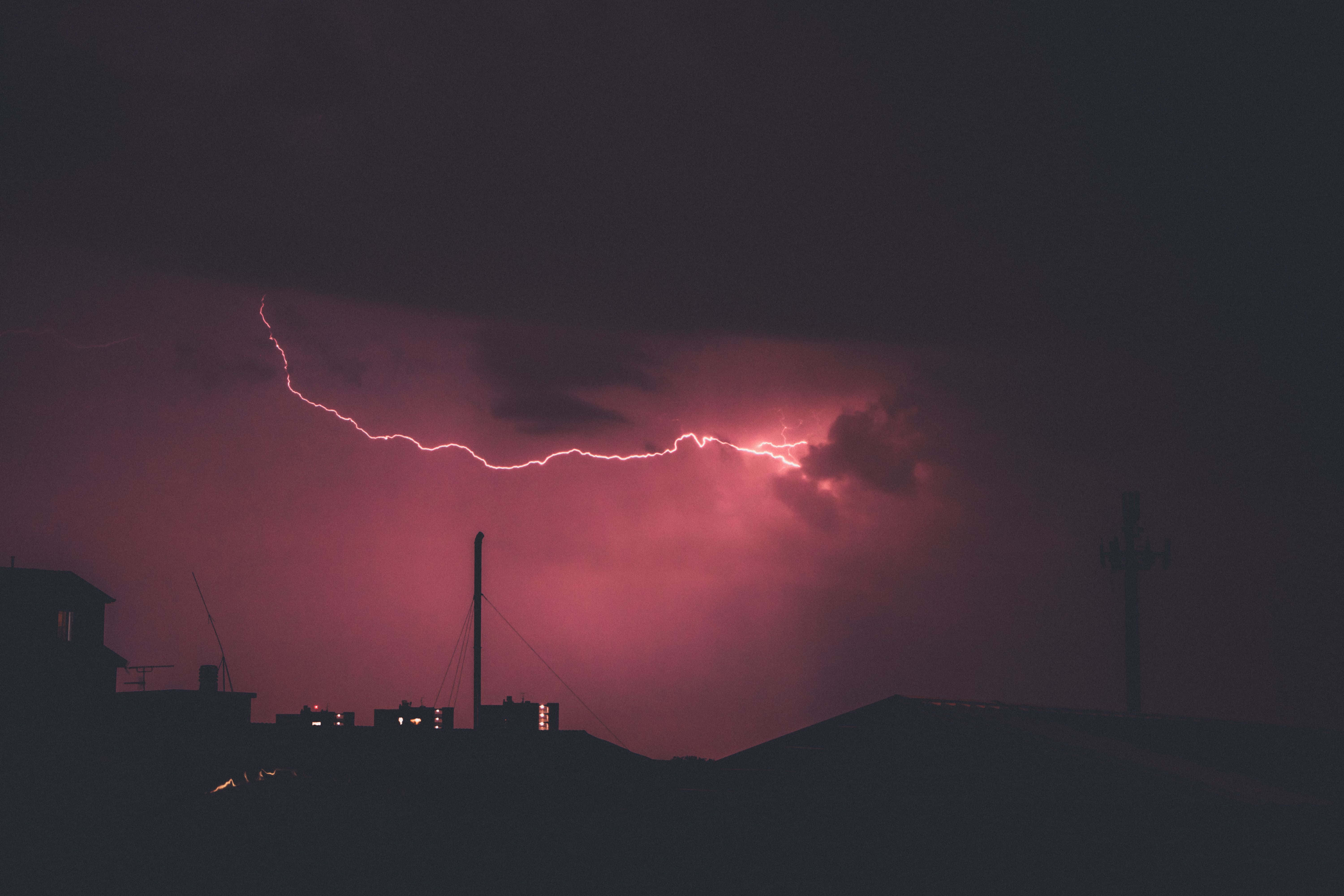 Red Lightning 桌布 壁紙 — 一道鮮豔的粉紅色閃電劃過暴風雨的夜空，映襯著屋頂和電線桿的剪影。