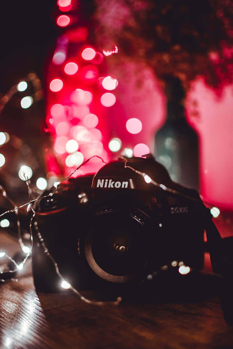 Dslr Camera 桌布 壁紙 — 一部 Nikon D5300 相機放置在木質表面上，夜晚被發光的紅色和白色仙女燈包圍。