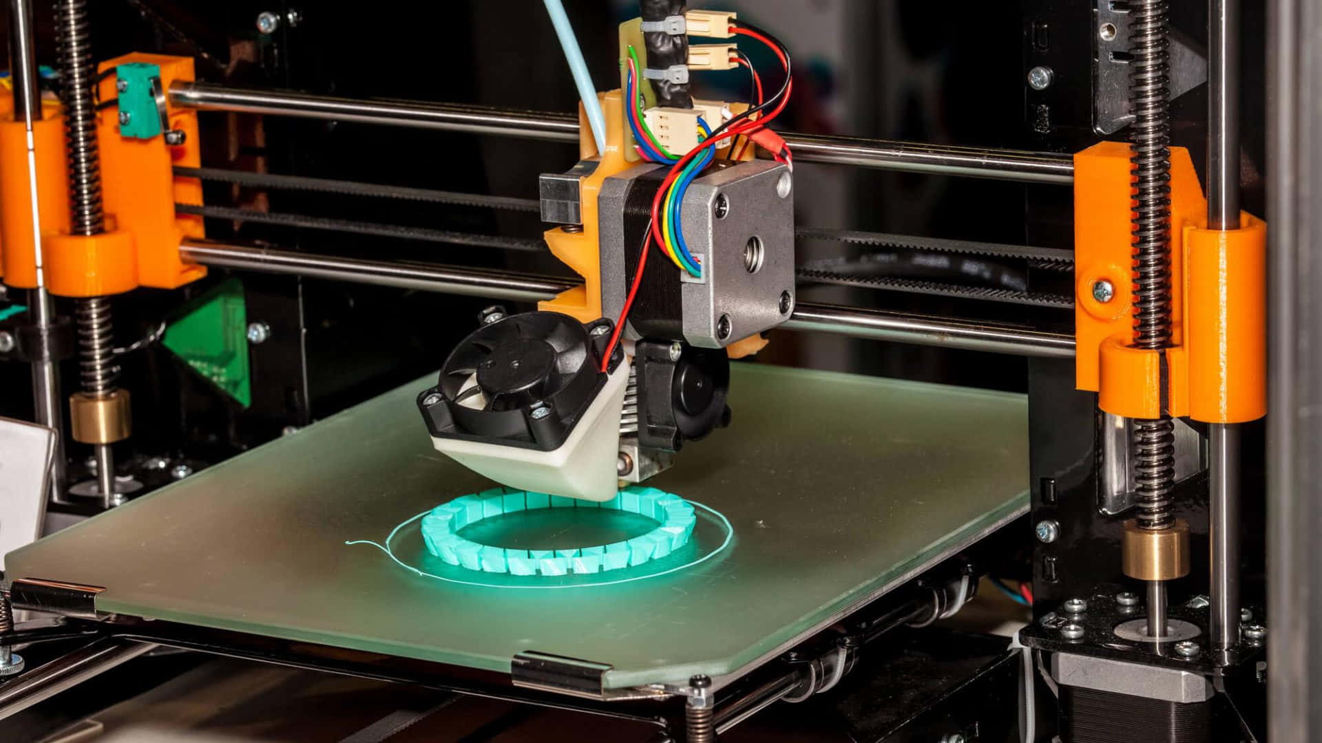 3d Printer 桌布 壁紙 — 一部3D打印機擠出發光的青綠色線材，在玻璃床上用橙色機械臂形成一個圓形齒輪。