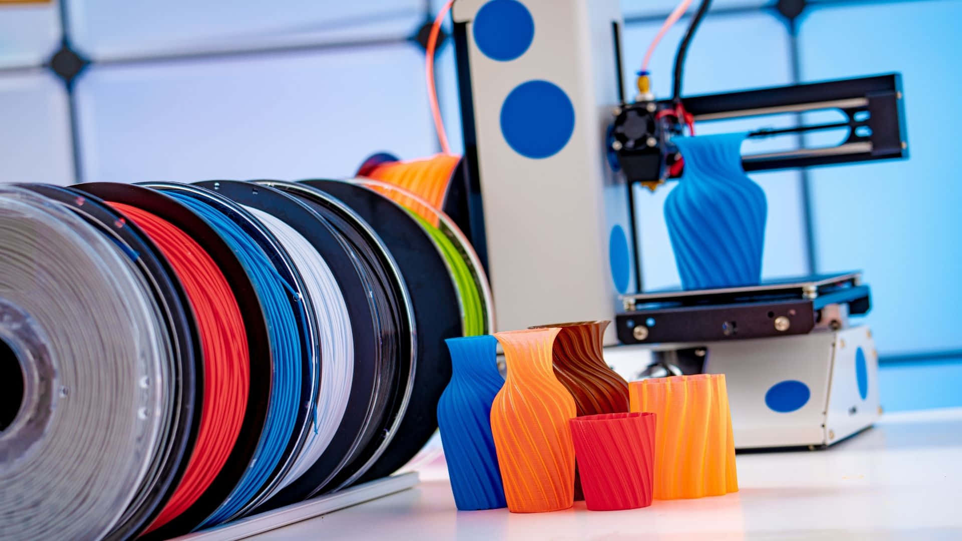 3d Printer 桌布 壁紙 — 一部3D打印機正在擠出藍色線材，而色彩繽紛的線軸和打印出來的花瓶則放在乾淨的白色工作枱面上。
