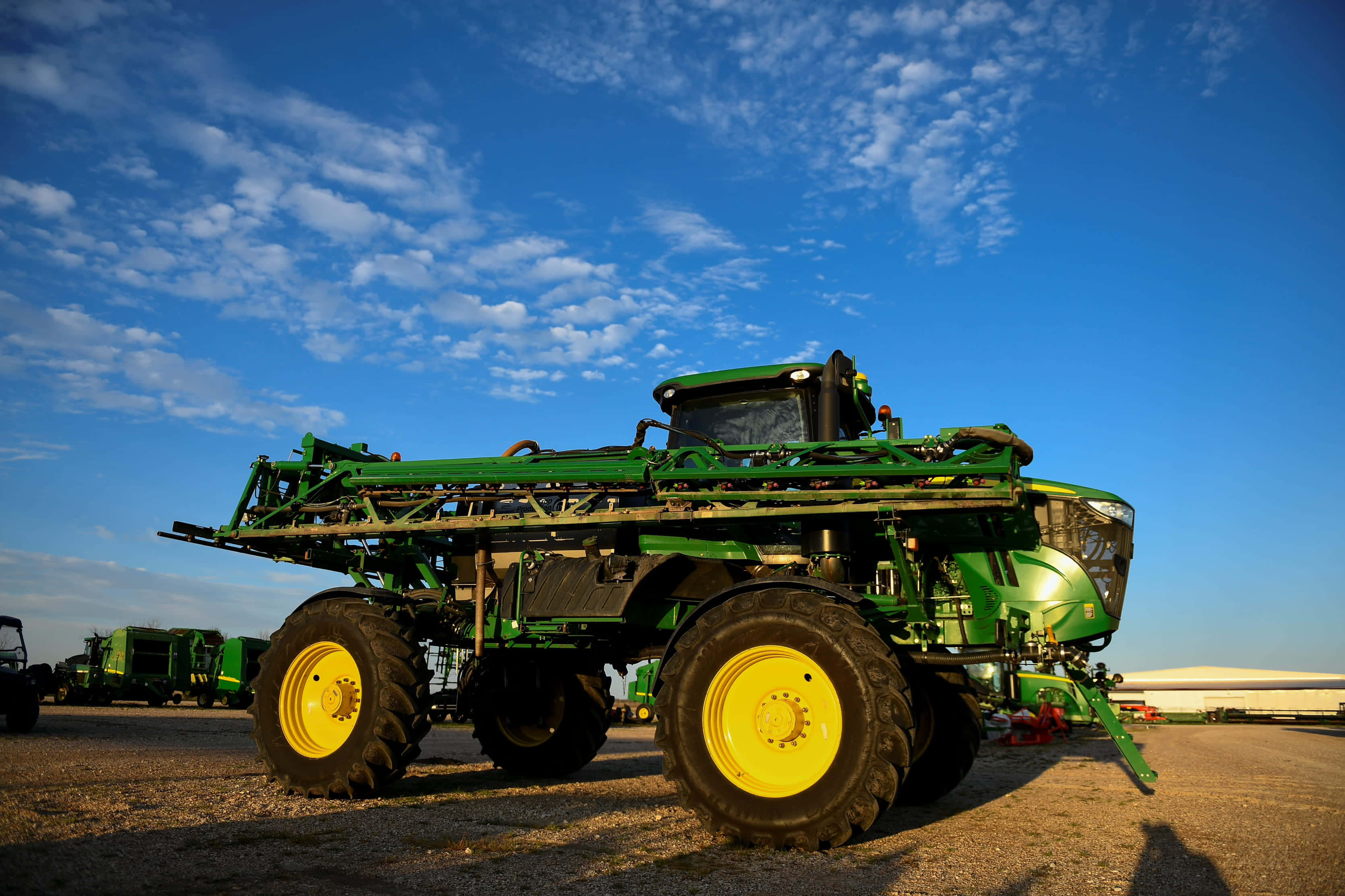 John Deere 桌布 壁紙 — 一部大型綠色John Deere噴霧器，配有黃色輪子，站在晴朗藍天和零星白雲下的碎石地上。