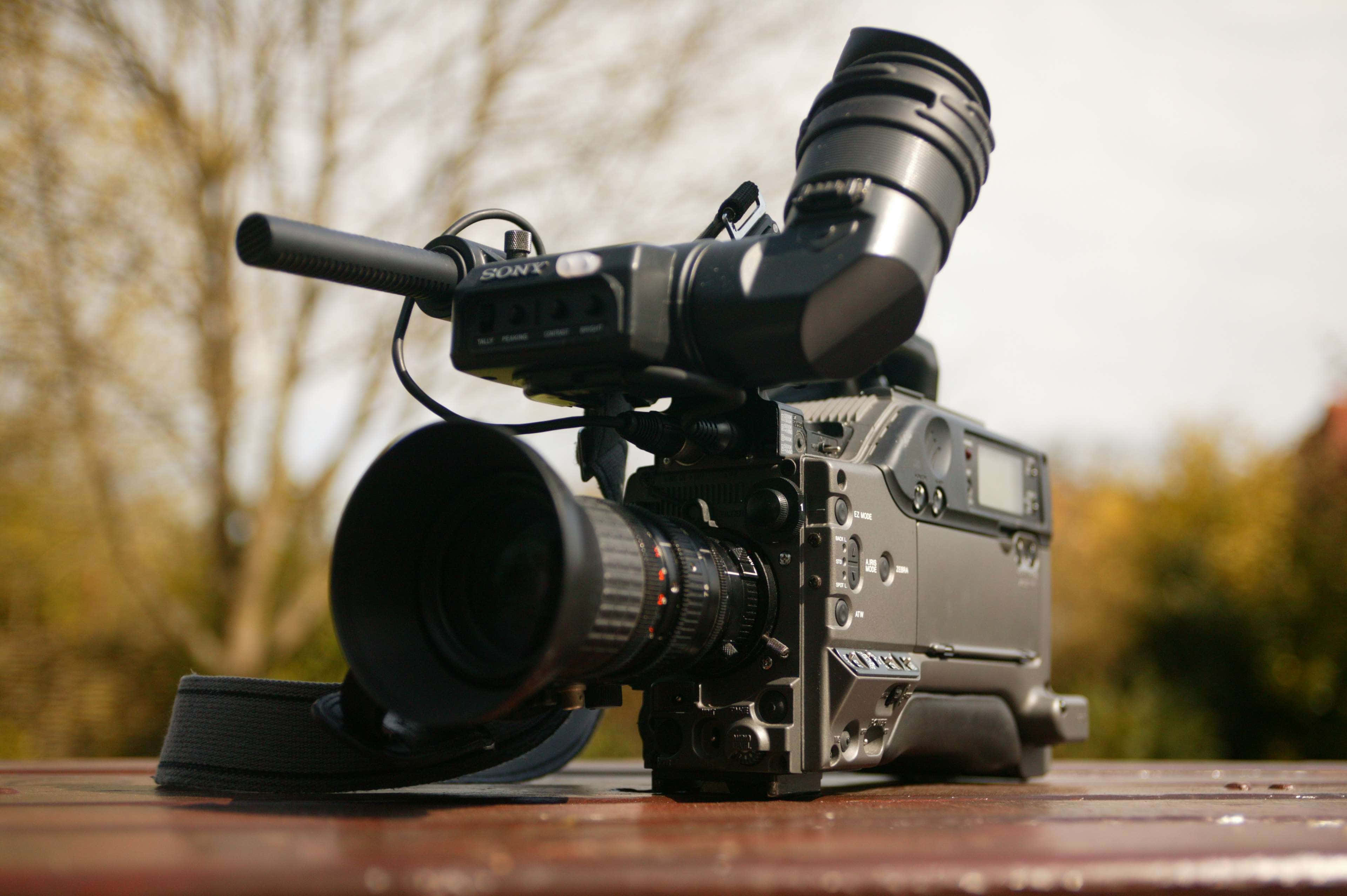 Broadcast 桌布 壁紙 — 一部專業Sony攝錄機，配備大型鏡頭和麥克風，放置在戶外木質表面上，背景是模糊的秋樹。