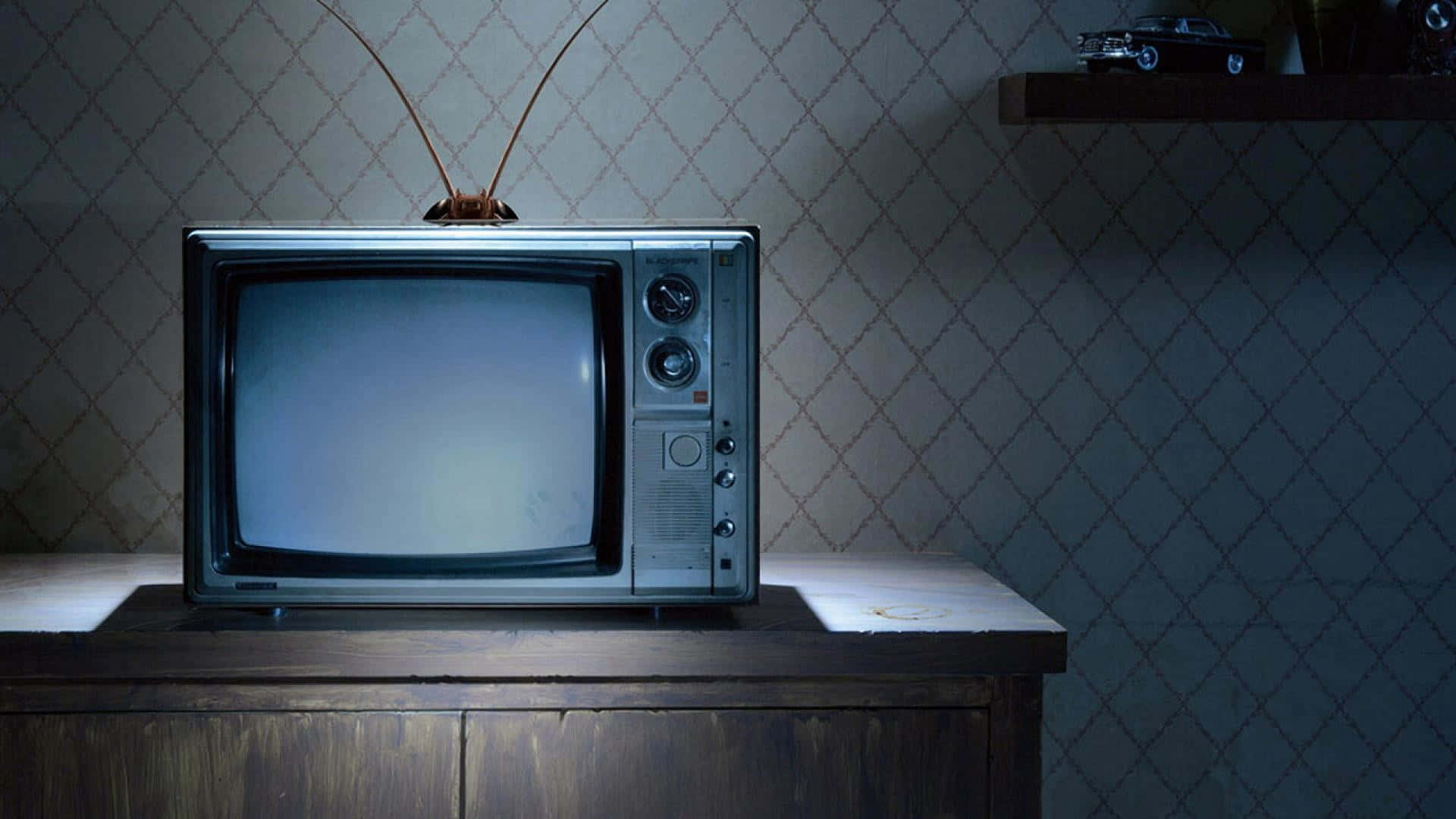 tv 桌布 壁紙 — 一部帶兔耳天線的復古銀色電視機，放在一個光線昏暗、貼有圖案牆紙的房間裡的木櫃上。