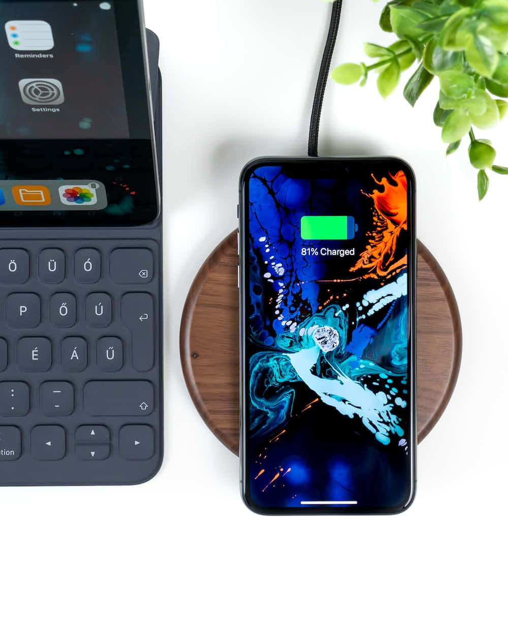 Wireless Charging 桌布 壁紙 — 一部帶有鮮豔藍色和橙色抽象壁紙的智能手機正在木質充電板上充電，旁邊有一個深灰色鍵盤和一株綠色植物。