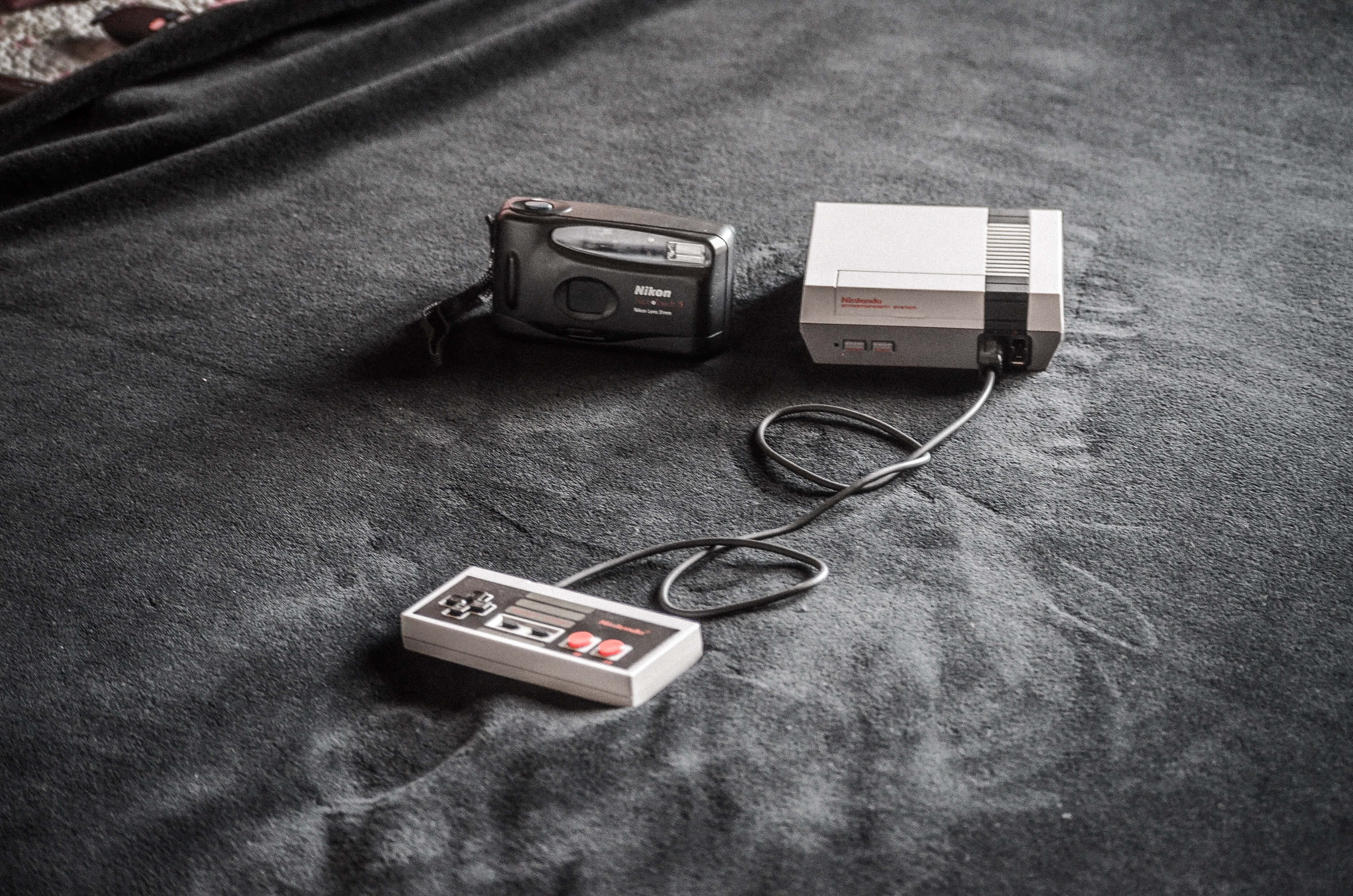 Gaming Consoles 桌布 壁紙 — 一部復古任天堂娛樂系統主機和控制器，與一部輕便數碼相機一同放在深色紋理布料表面上。