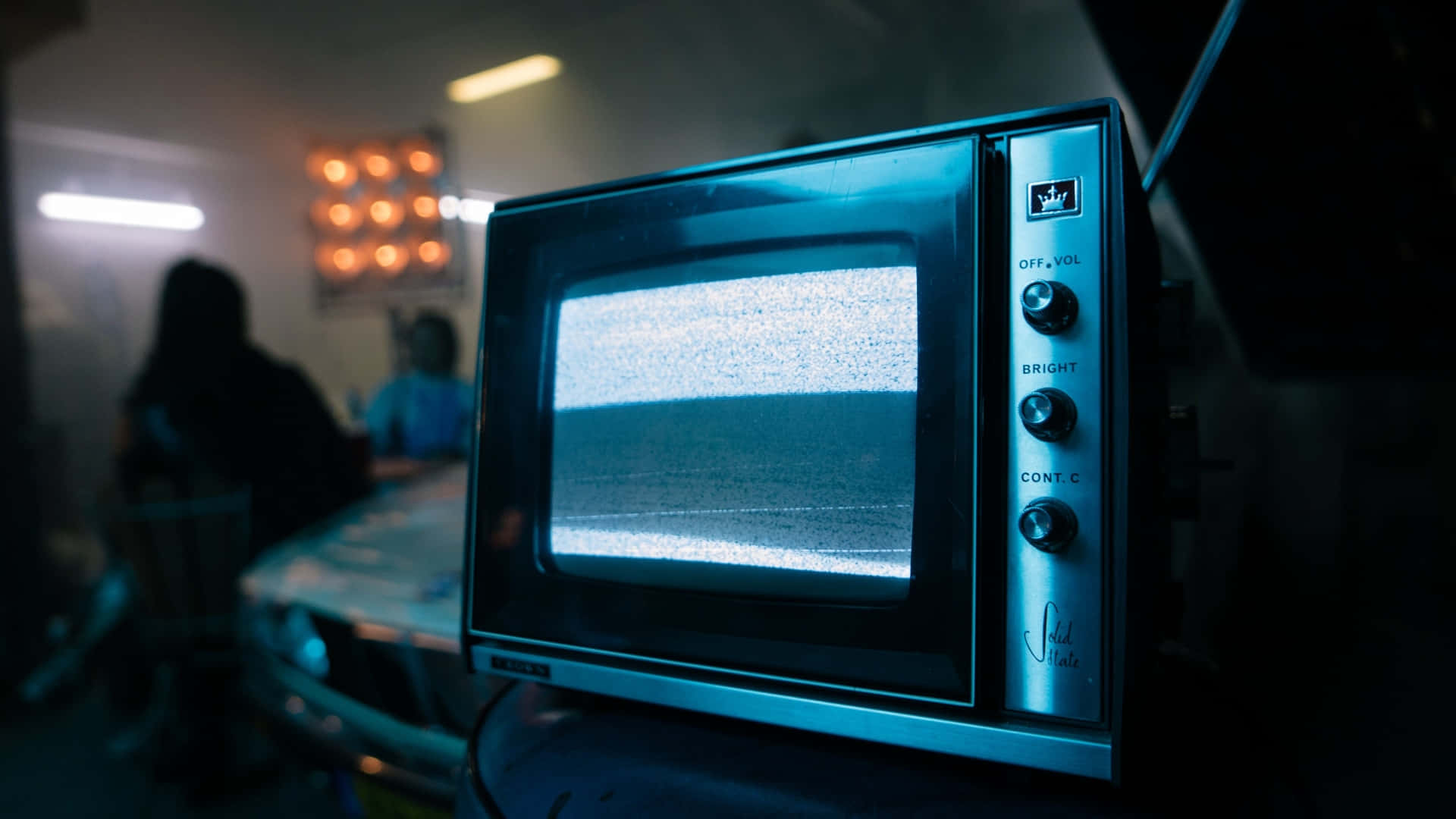Television Set 桌布 壁紙 — 一部復古電視機屏幕上顯示著雪花靜態畫面，放置在光線昏暗的房間中，背景有模糊的人影。