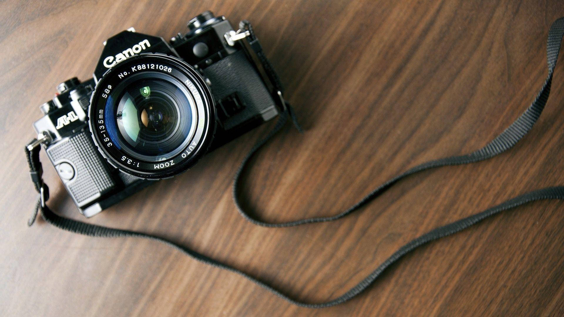 Dslr Camera 桌布 壁紙 — 一部復古黑色佳能相機配備變焦鏡頭，放置在溫暖的木質表面上，相機帶垂掛在附近。