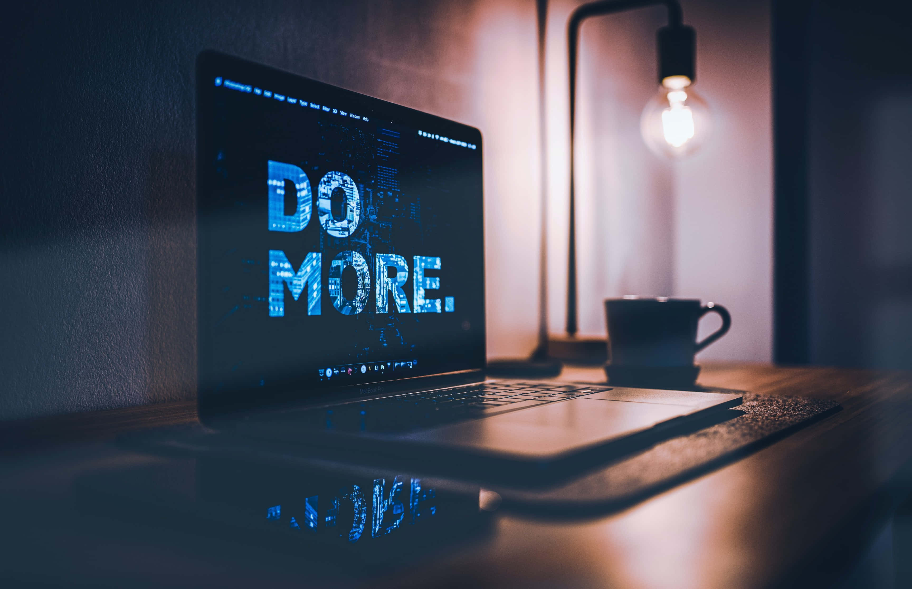 macbook pro 桌布 壁紙 — 一部手提電腦放在木桌上，以發光的藍色文字顯示「DO MORE.」，而燈泡的溫暖光線照亮了附近的咖啡杯。