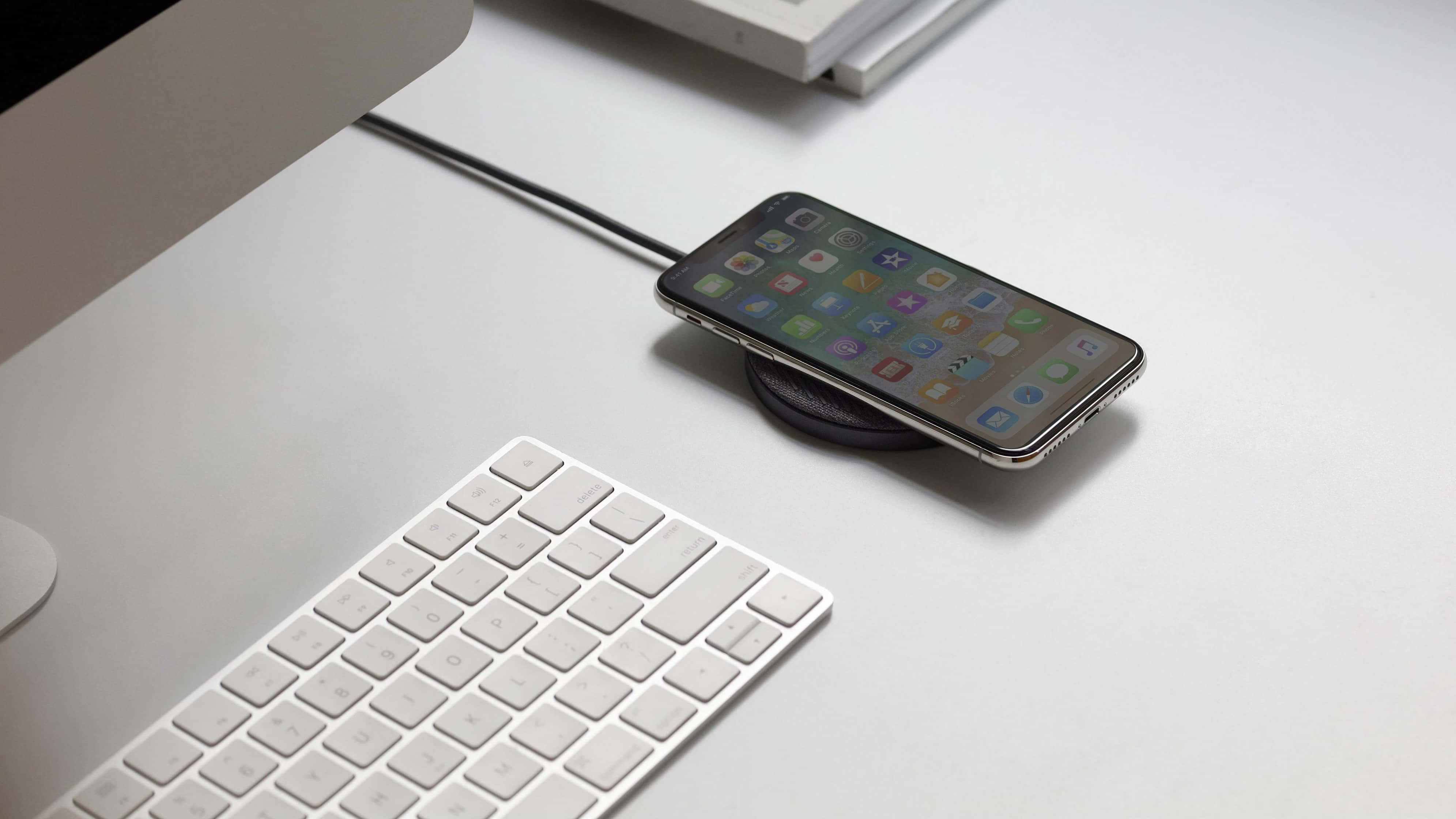 Wireless Charging 桌布 壁紙 — 一部時尚智能手機放在無線充電器上，旁邊是白色鍵盤和顯示器，置於簡約白色桌上。
