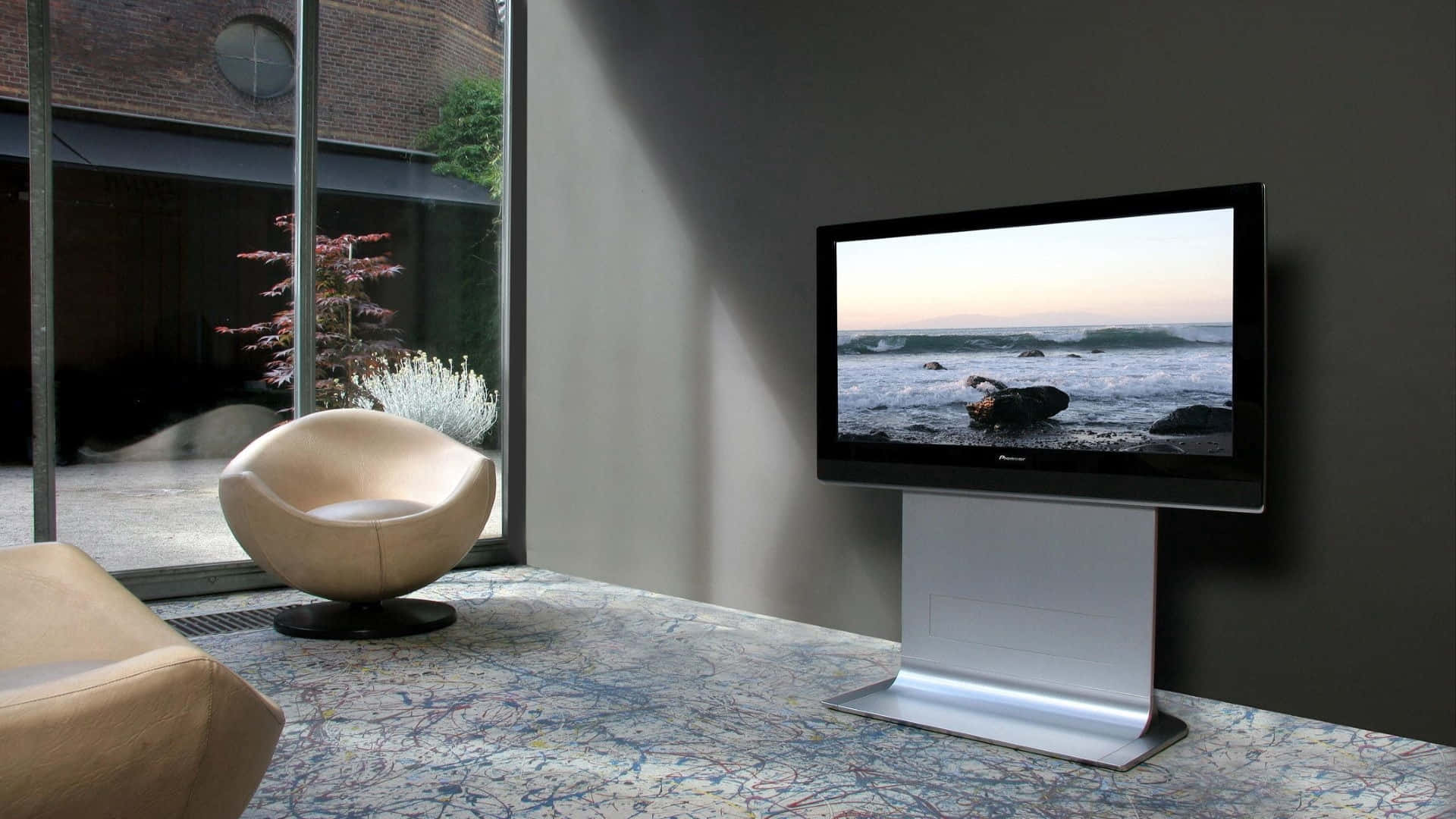 Television Set 桌布 壁紙 — 一部時尚現代電視顯示寧靜的海洋日落，同時放置在圖案地毯上，旁邊是一張弧形米色椅子，位於簡約的起居空間中。