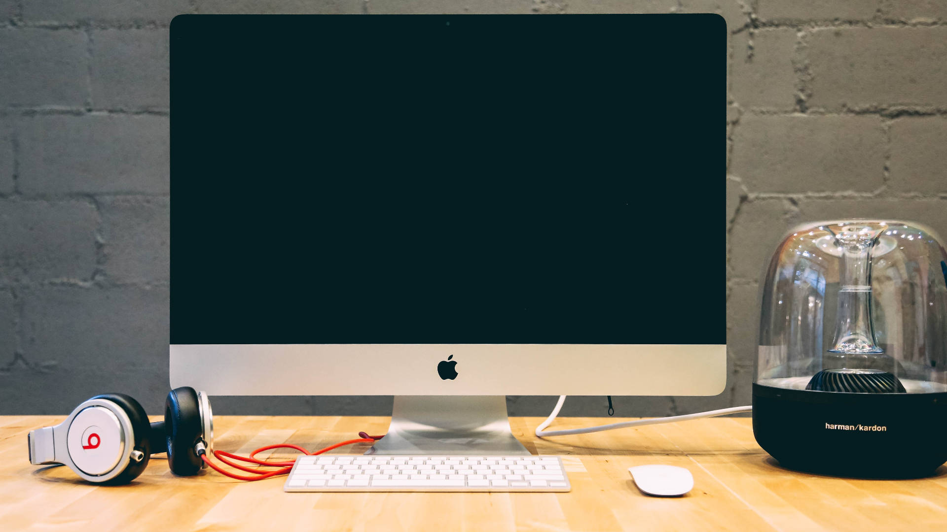 Mac 桌布 壁紙 — 一部時尚的Apple iMac放在木桌上，旁邊有Beats耳機和Harman Kardon揚聲器，背景是灰色磚牆。