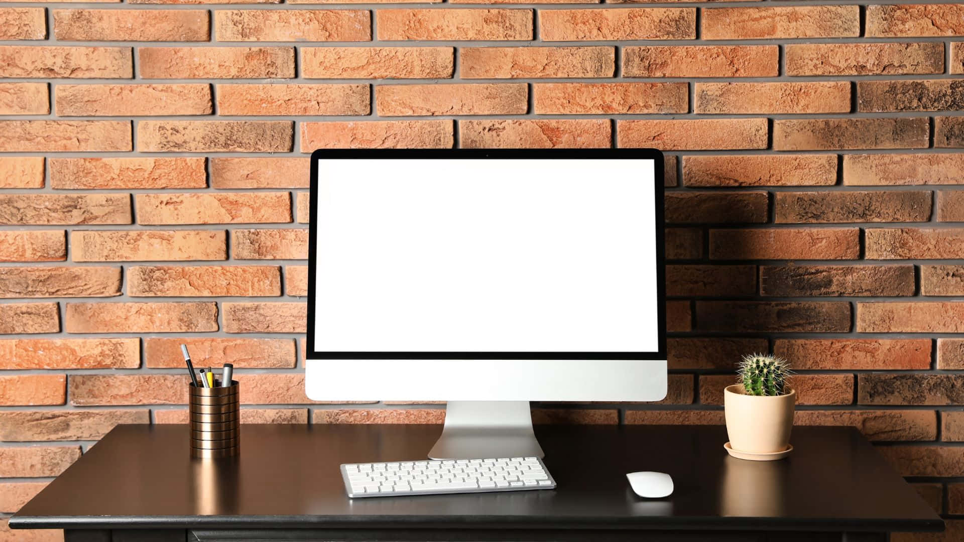 Computer Screen 桌布 壁紙 — 一部時尚的桌上電腦放在深色木桌上，背景是溫暖的磚牆，附近有一個小仙人掌和筆筒。