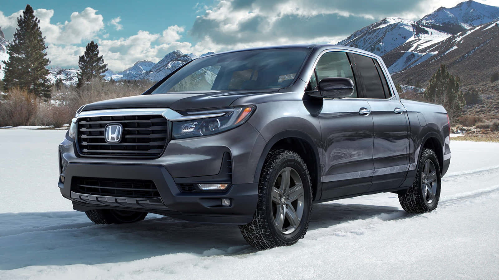Honda Ridgeline 桌布 壁紙 — 一部時尚的灰色本田Ridgeline皮卡車停在雪地上，背景是積雪的山脈和常青樹，天空部分多雲。