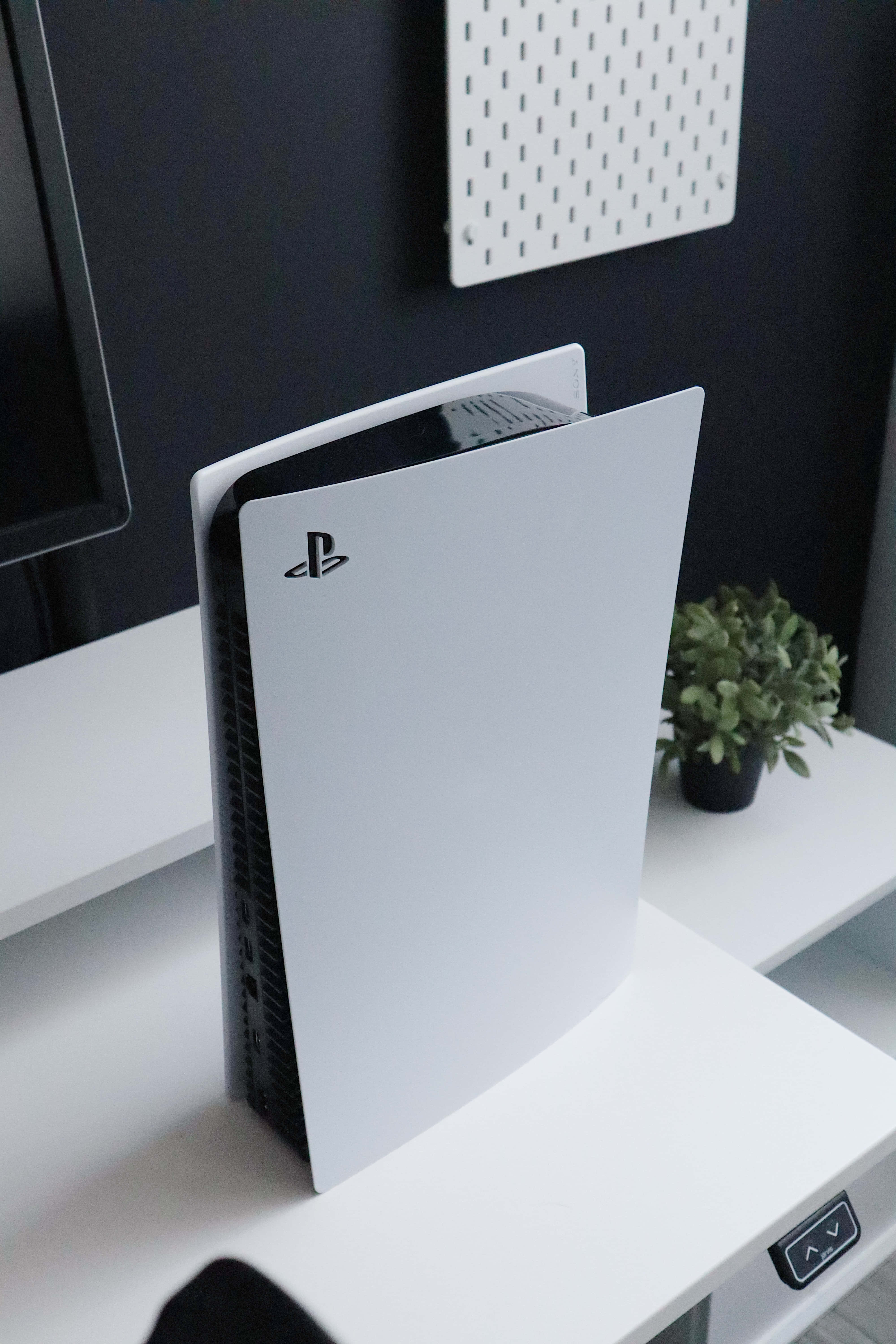 Gaming Consoles 桌布 壁紙 — 一部時尚的白色 PlayStation 5 主機直立放置在簡約的白色桌子上，旁邊有一棵小綠色植物和一個顯示器。