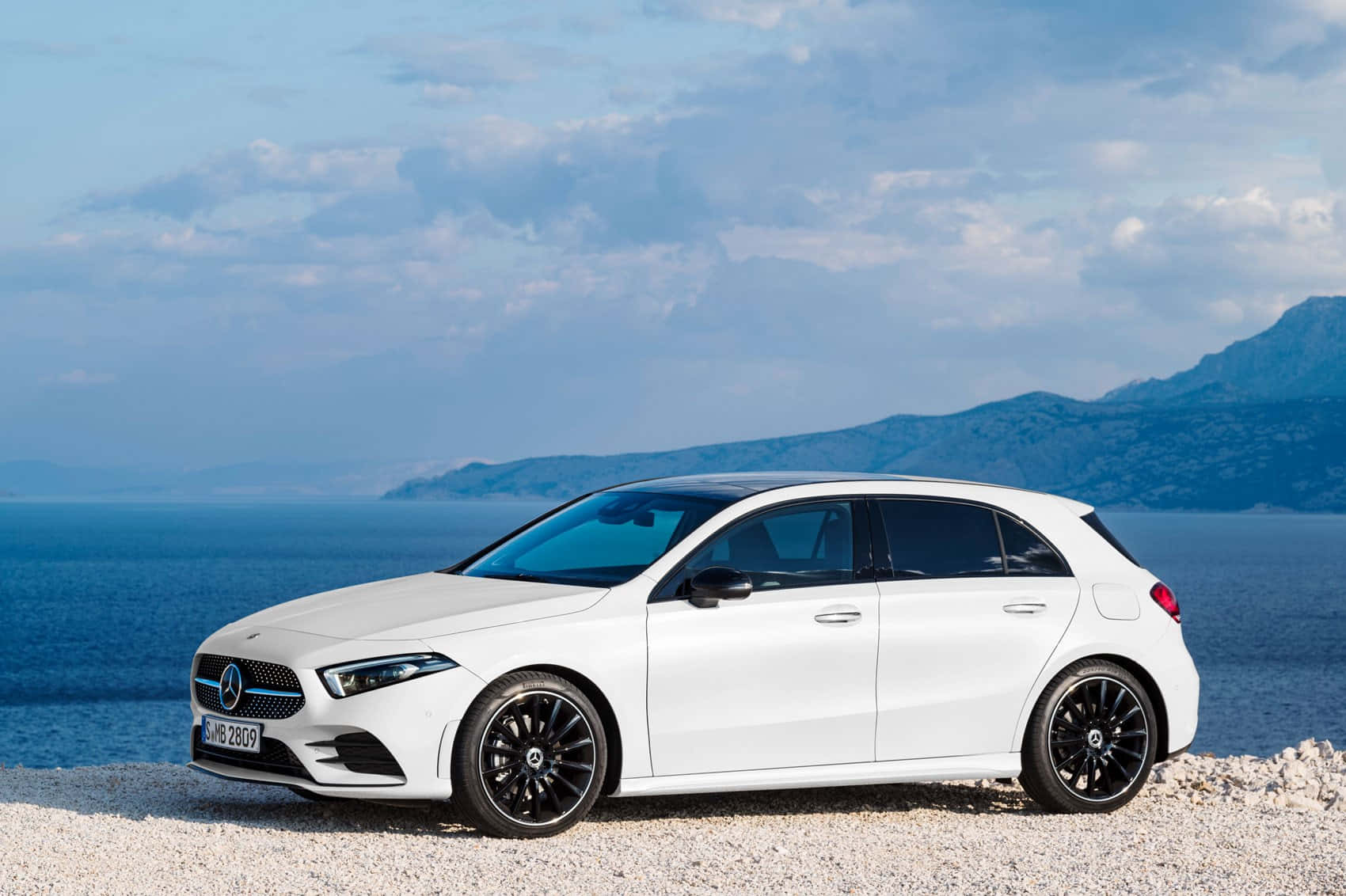 Mercedes Benz A-class 桌布 壁紙 — 一部時尚的白色平治A-Class掀背車停泊在卵石灘上，背景是廣闊的藍色大海和遠山，天空部分多雲。