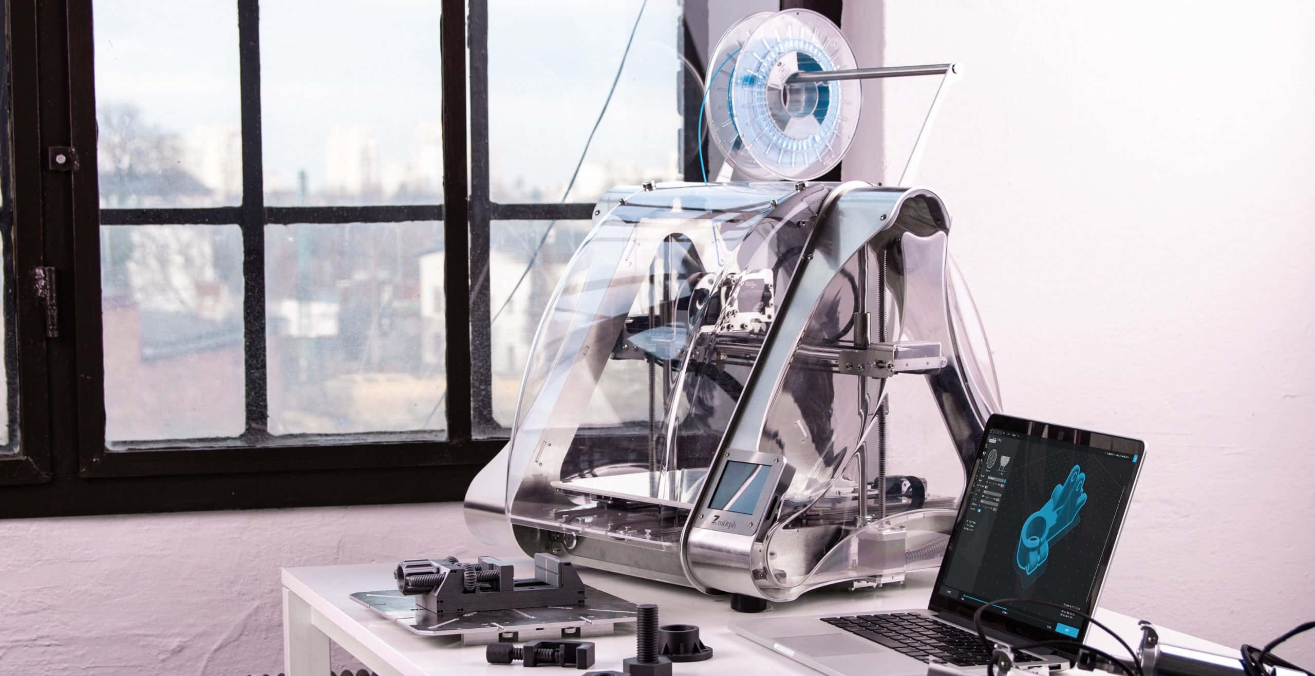 3d Printer 桌布 壁紙 — 一部時尚的透明3D打印機放在白色桌子上，旁邊是一部顯示藍色3D模型的筆記本電腦，透過大窗戶可以看到城市景觀。
