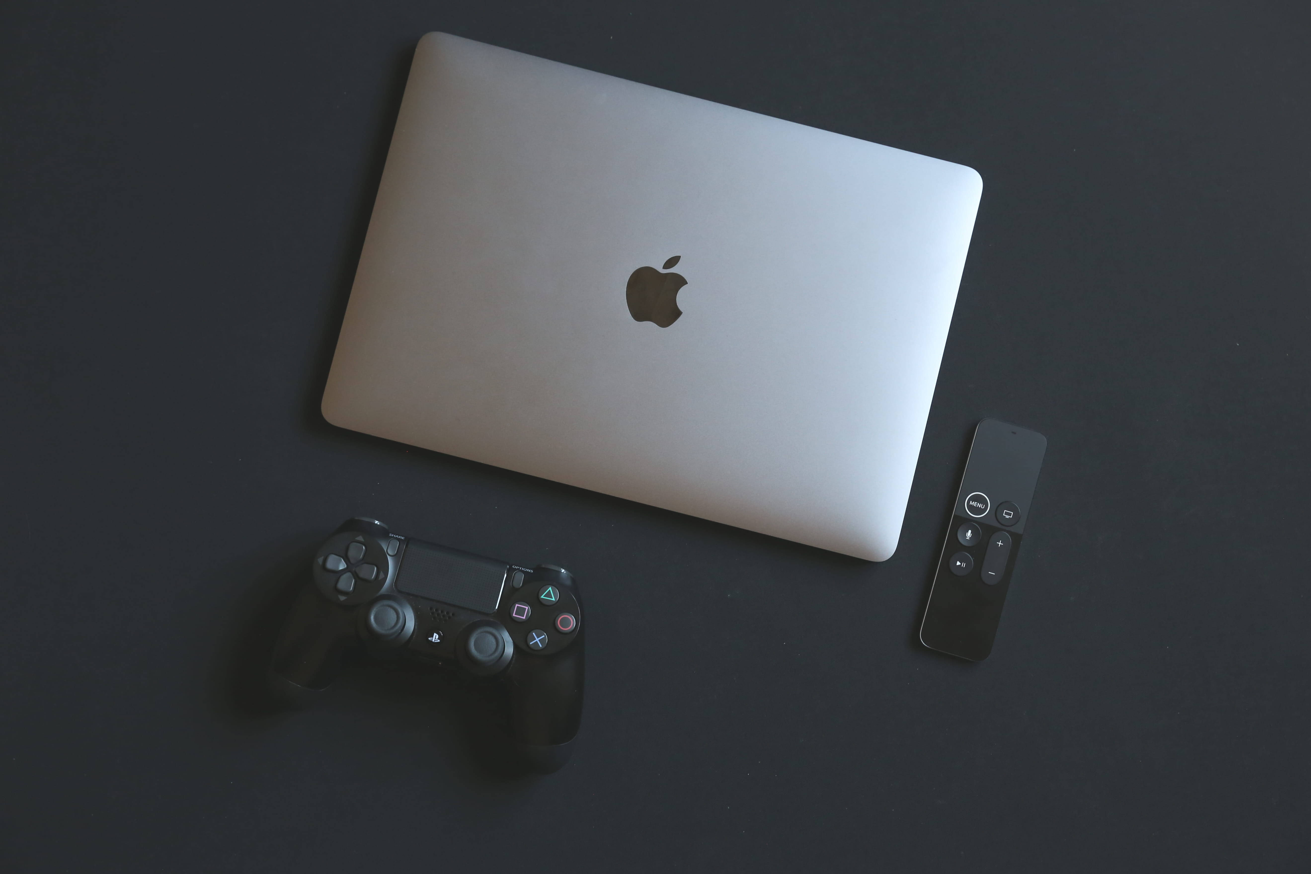  壁紙 — 一部時尚的銀色 MacBook 手提電腦旁邊放著一個黑色 PlayStation 控制器和 Apple TV 遙控器，置於深色表面上。