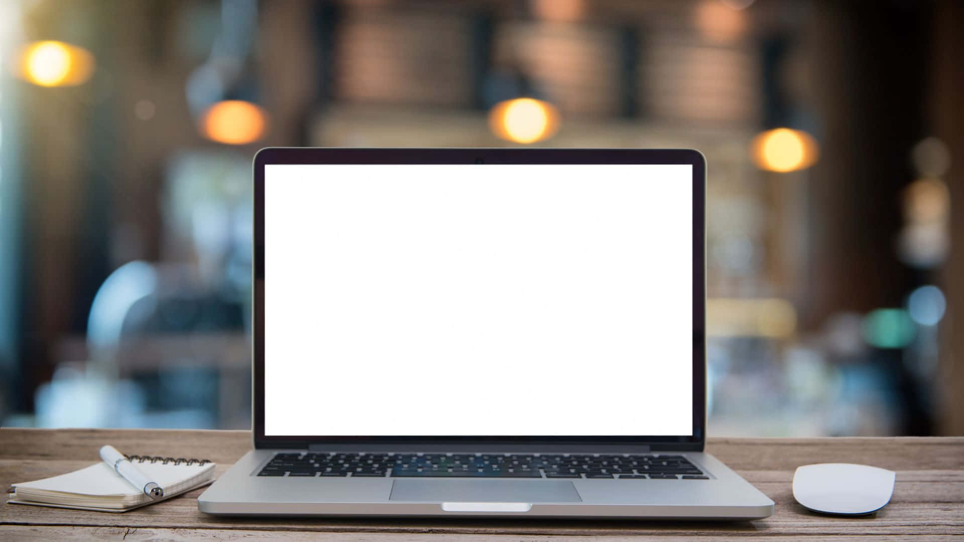Computer Screen 桌布 壁紙 — 一部時尚的銀色手提電腦，配備空白白色屏幕，放在木桌上，旁邊有筆記本和滑鼠，位於燈光溫暖的咖啡館內。