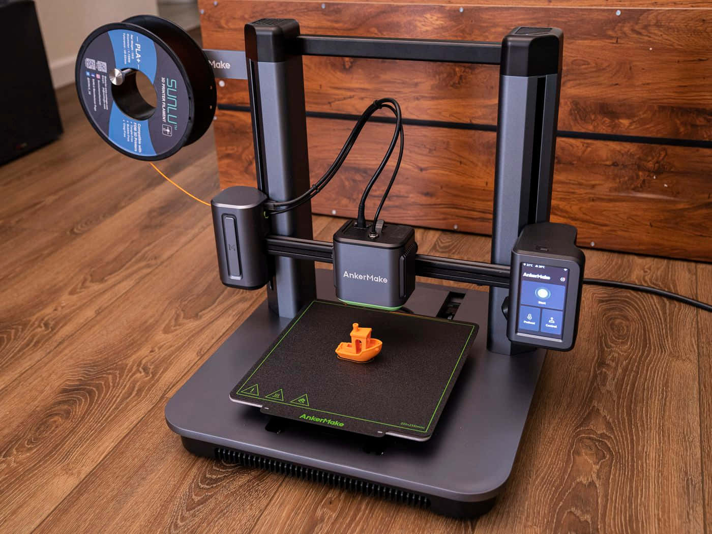 3d Printer 桌布 壁紙 — 一部時尚的黑色AnkerMake 3D打印機放在木地板上，打印床上有一個小的橙色打印船，上方裝有線材卷。