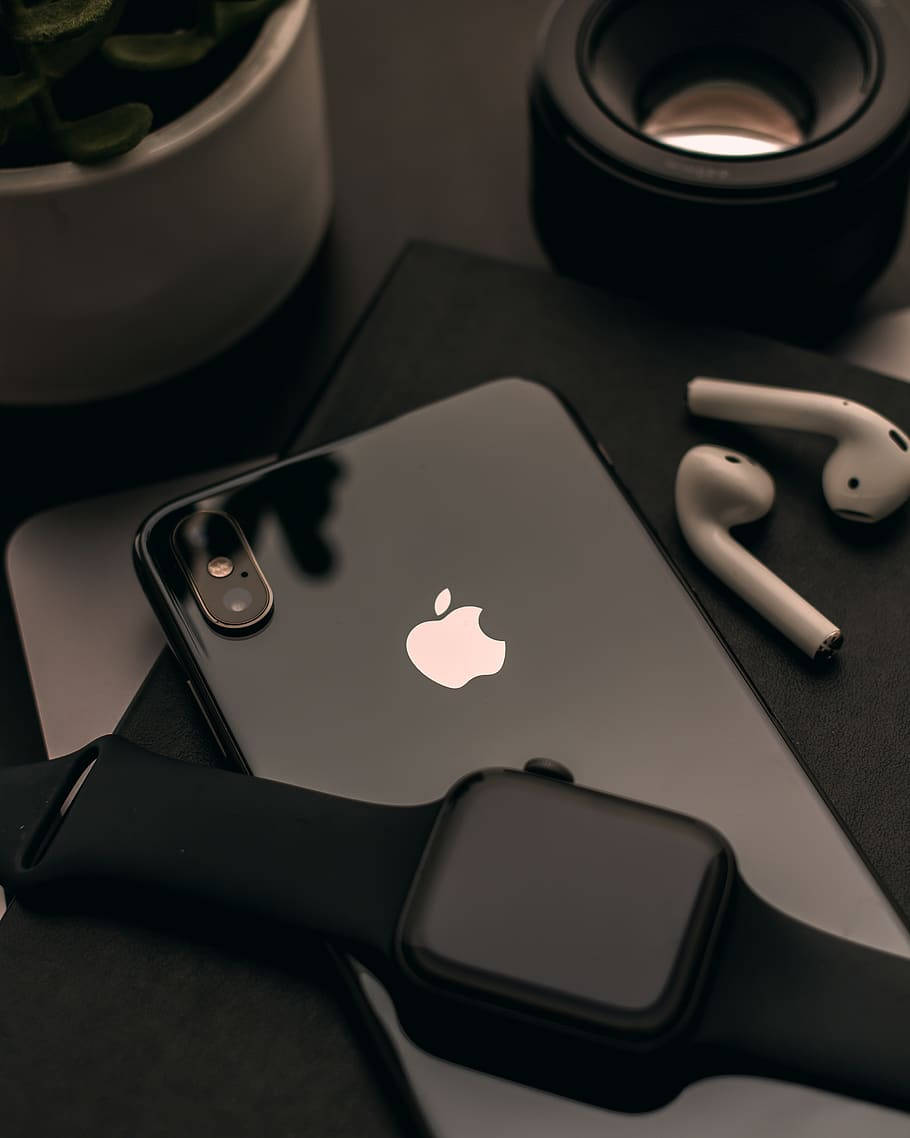 蘋果 桌布 壁紙 — 一部時尚的黑色iPhone和Apple Watch與白色AirPods和盆栽植物一起放在深色表面上。