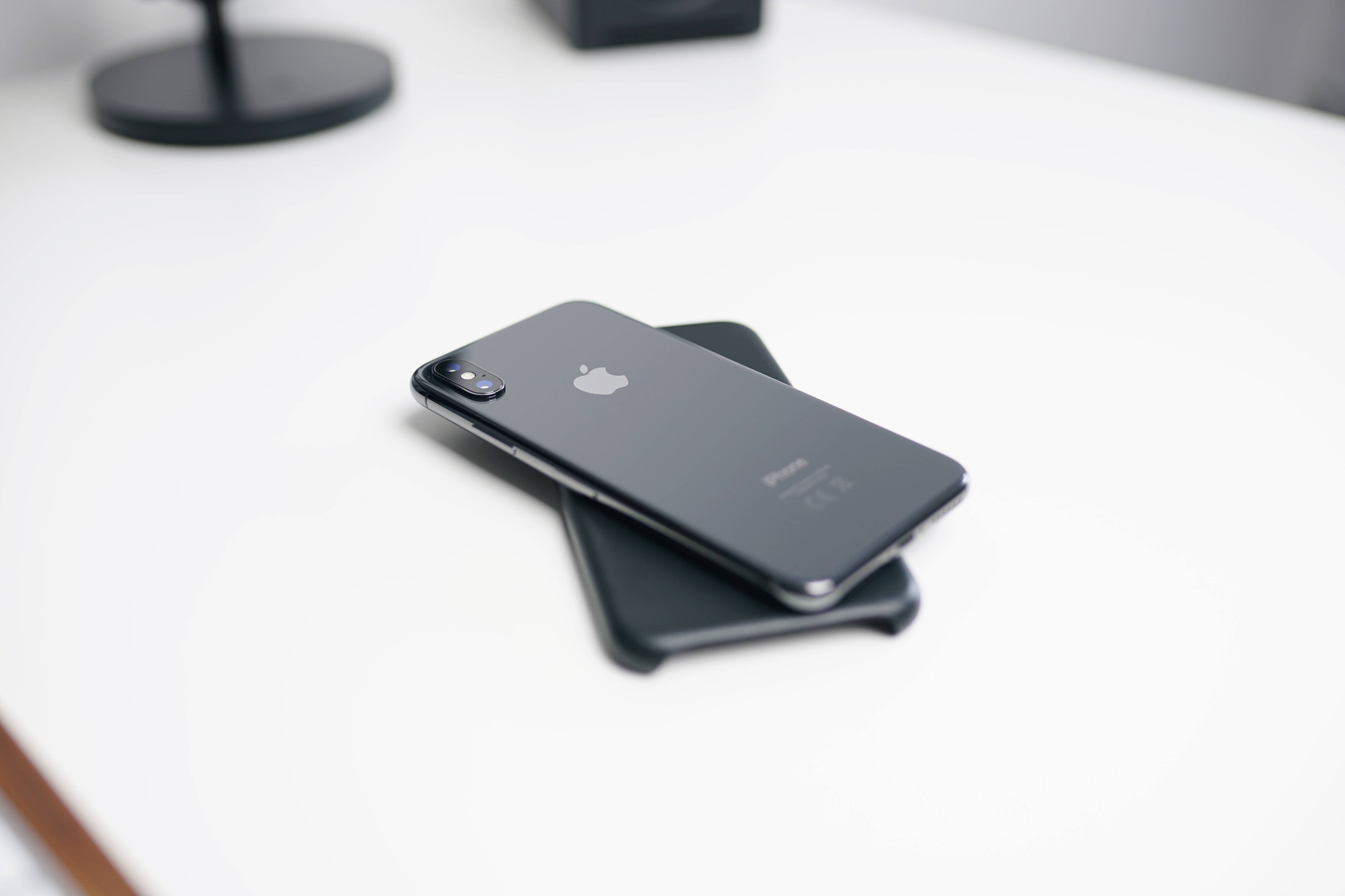 iPhone X iOS 13 壁紙 — 一部時尚的黑色iPhone放在另一部手機上，置於簡約白色桌面，帶有柔和陰影。