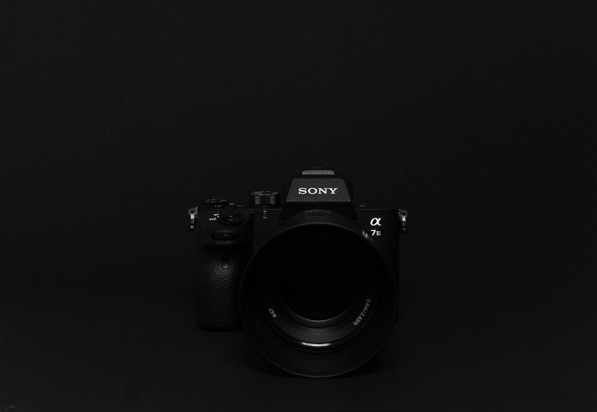 黑色動態圖 桌布 壁紙 — 一部時尚的黑色Sony Alpha 7 III相機，在極簡純黑背景下以柔和工作室燈光居中展示。