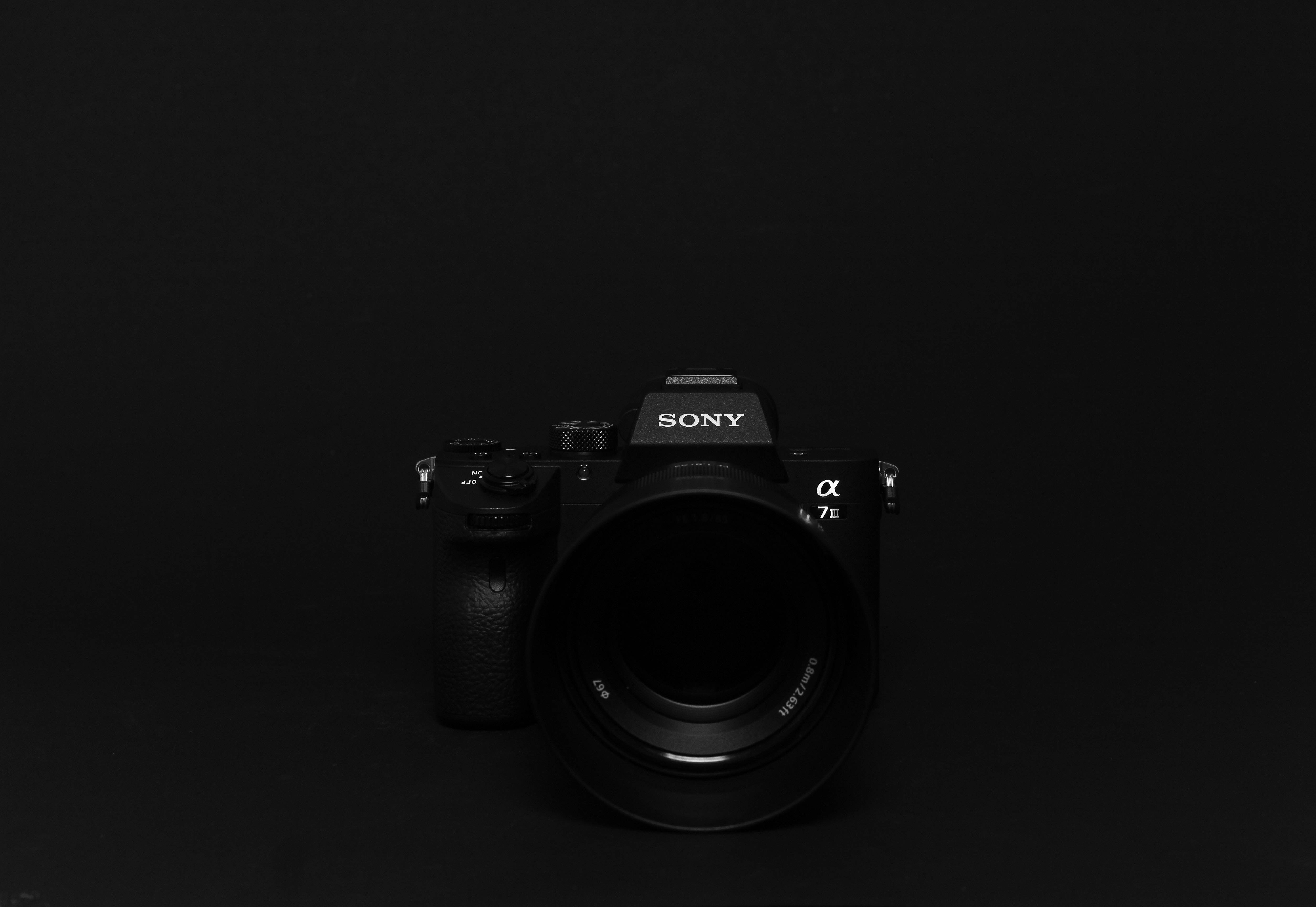 黑色動態圖 桌布 壁紙 — 一部時尚的黑色Sony Alpha 7 III相機，在極簡純黑背景上居中，配有柔和的工作室燈光。