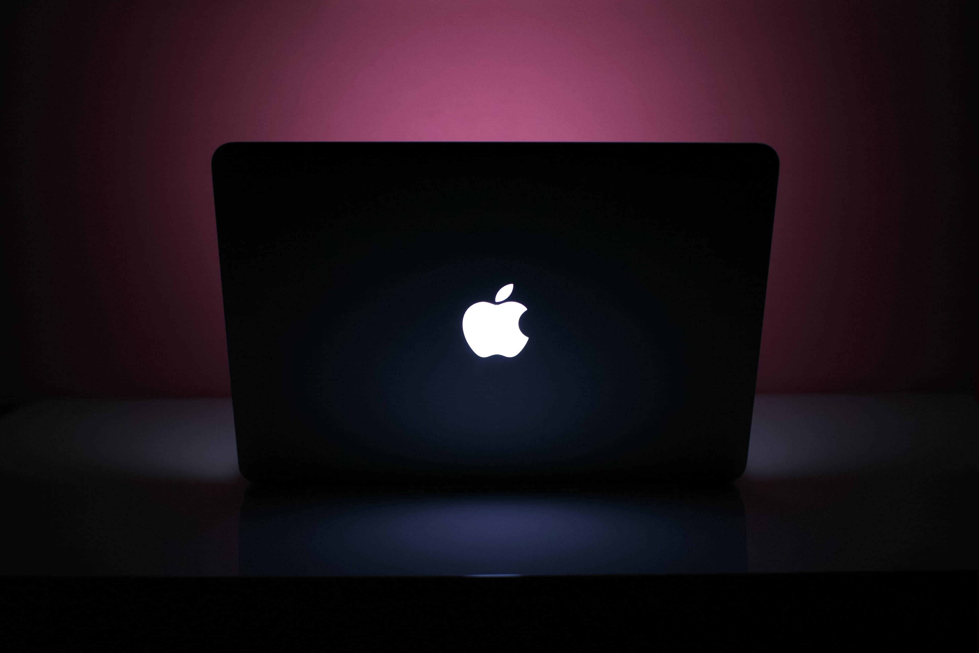 MacBook Pro 桌布 壁紙 — 一部時尚的黑色手提電腦，帶有發光的白色Apple標誌，放置在柔和粉紅色環境照明下的深色表面上。