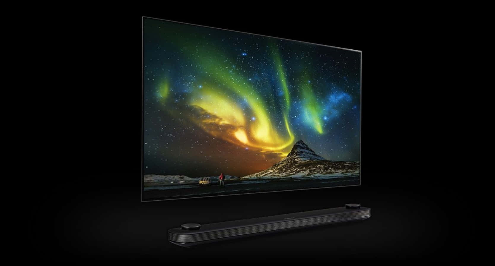 tv 桌布 壁紙 — 一部時尚的黑色電視顯示出充滿活力的北極光在雪山上方舞動，下方有兩個小人影在觀看。