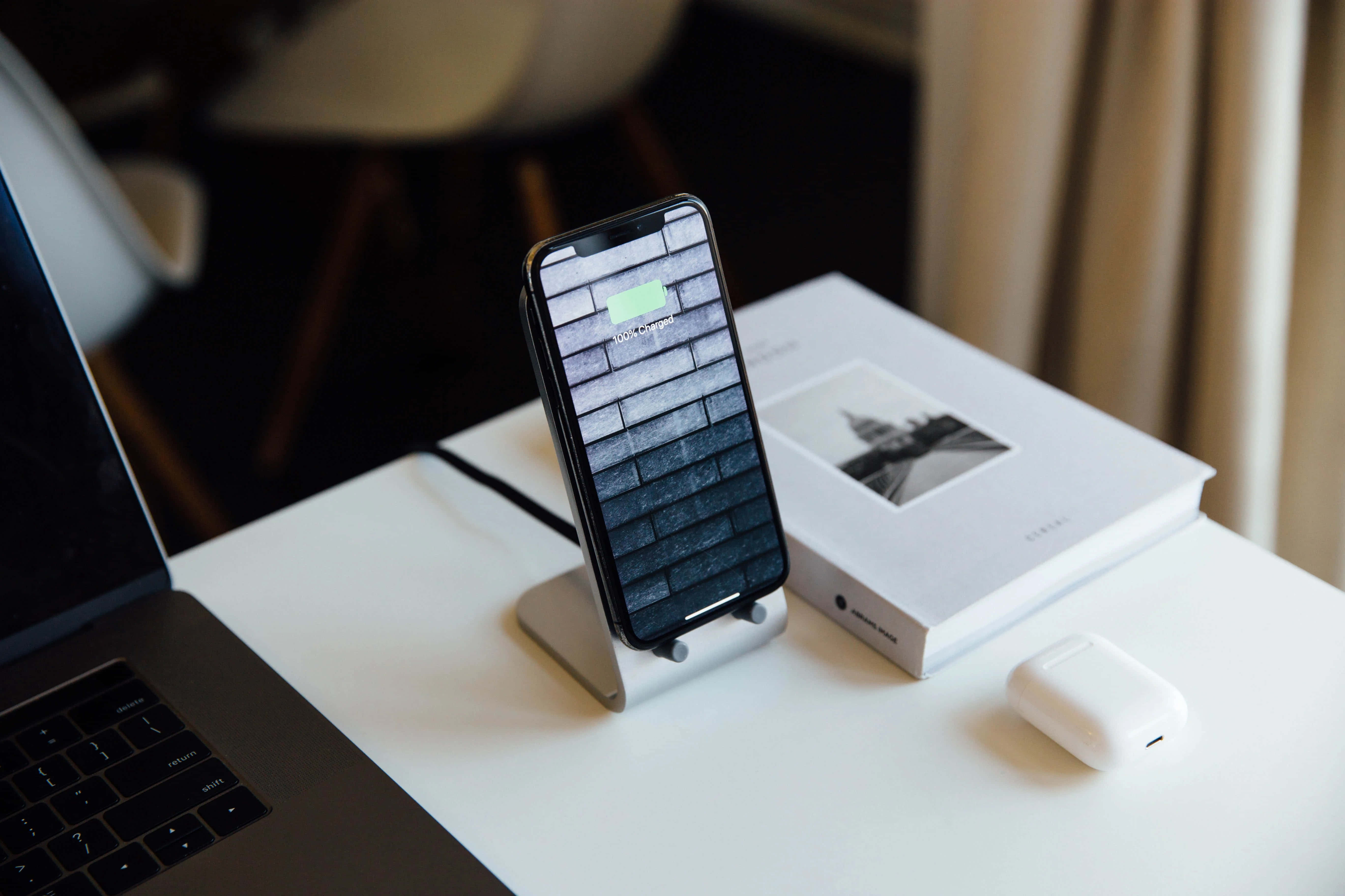 Wireless Charging 桌布 壁紙 — 一部智能手機在支架上充電，旁邊有一部手提電腦和AirPods，放在簡約的白色桌子上。