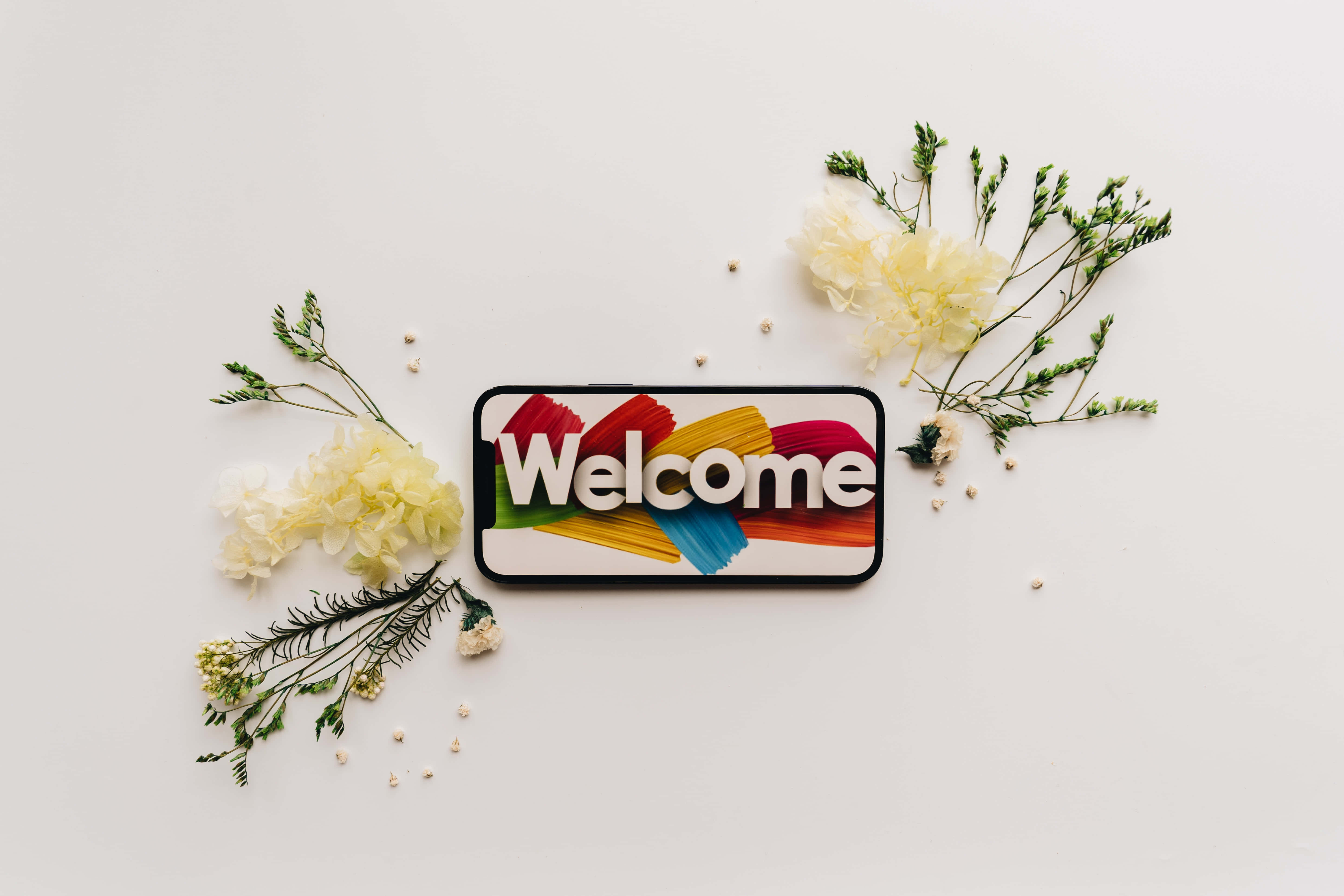 Welcome 桌布 壁紙 — 一部智能手機在白色表面上顯示色彩繽紛的「歡迎」圖案，周圍環繞著精緻的黃色花朵和綠色植物。