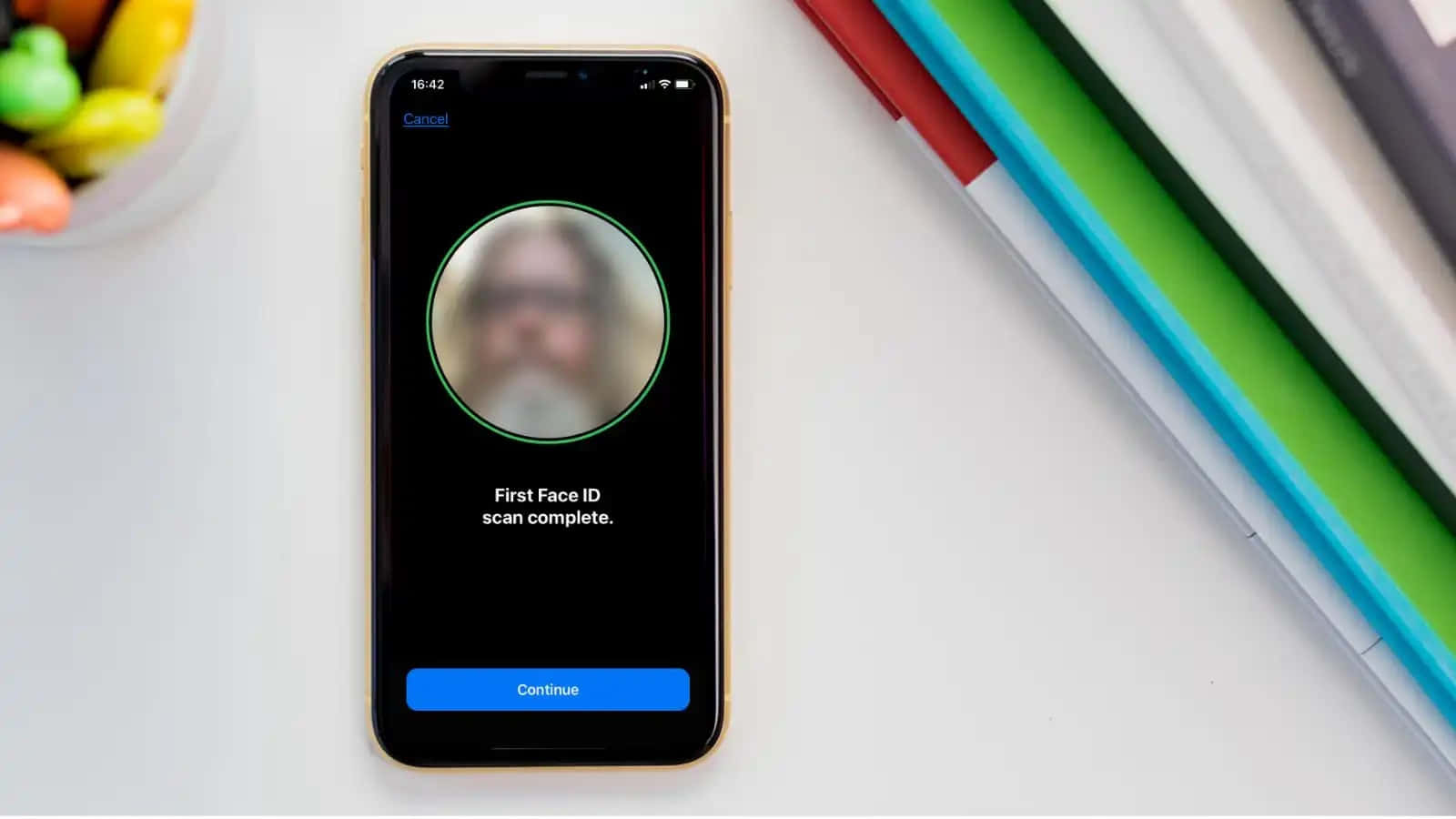 Face Id 桌布 壁紙 — 一部智能手機顯示已完成的 Face ID 掃描，附近有模糊的臉孔和彩色筆在白色桌子上。