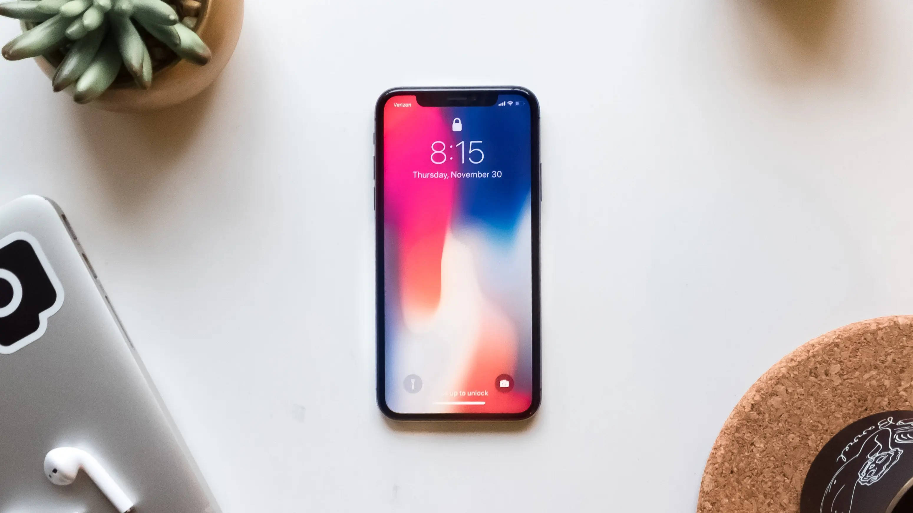 一部有趣的iPhone，讓你從人群中脫穎而出 壁紙 — 一部顯示彩色漸變桌布的智能手機放在白色書桌中央，旁邊有一盆多肉植物和耳塞。