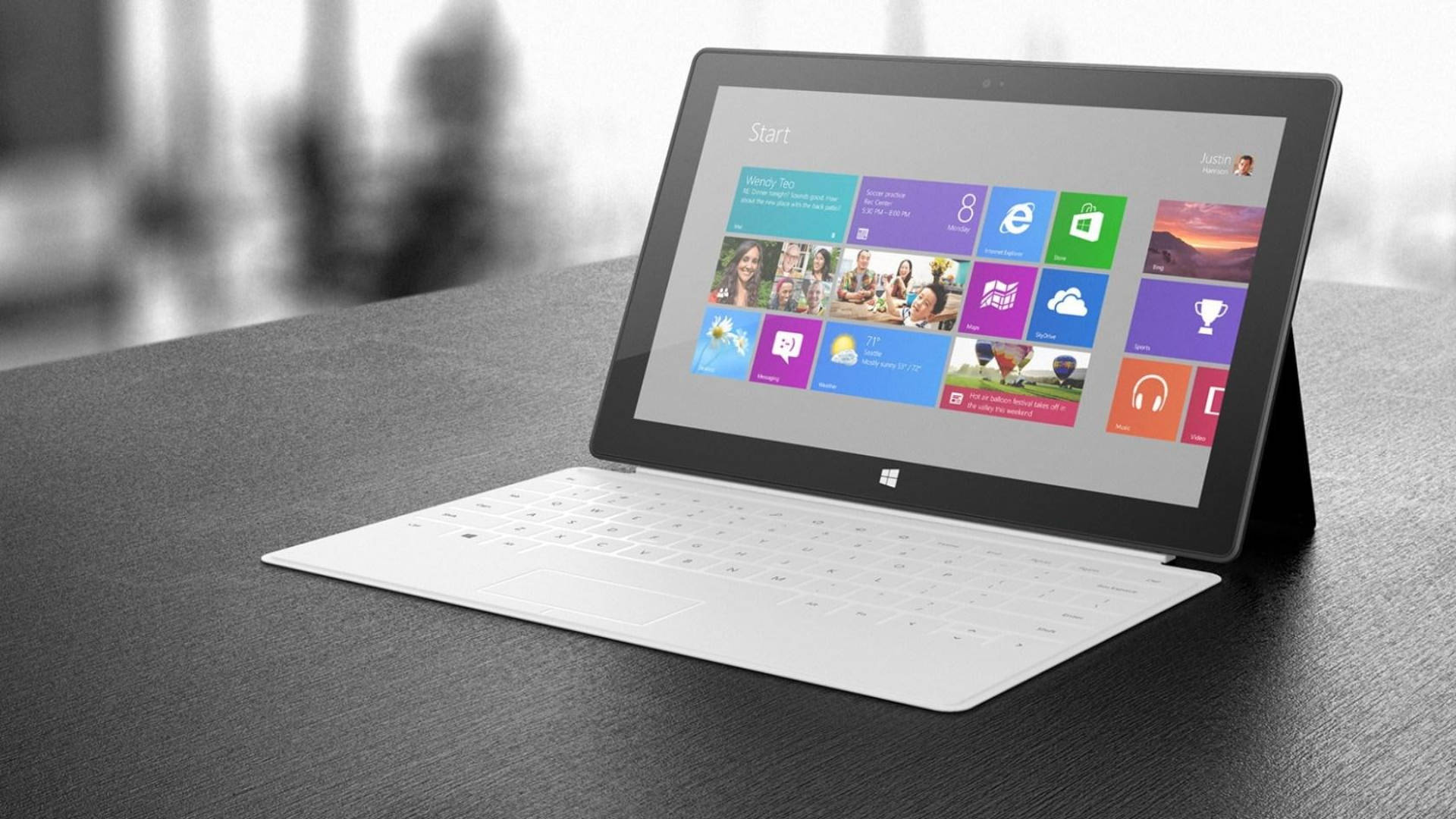 microsoft 桌布 壁紙 — 一部白色Microsoft Surface平板電腦配備鍵盤保護蓋，在深色紋理桌上顯示Windows開始畫面。