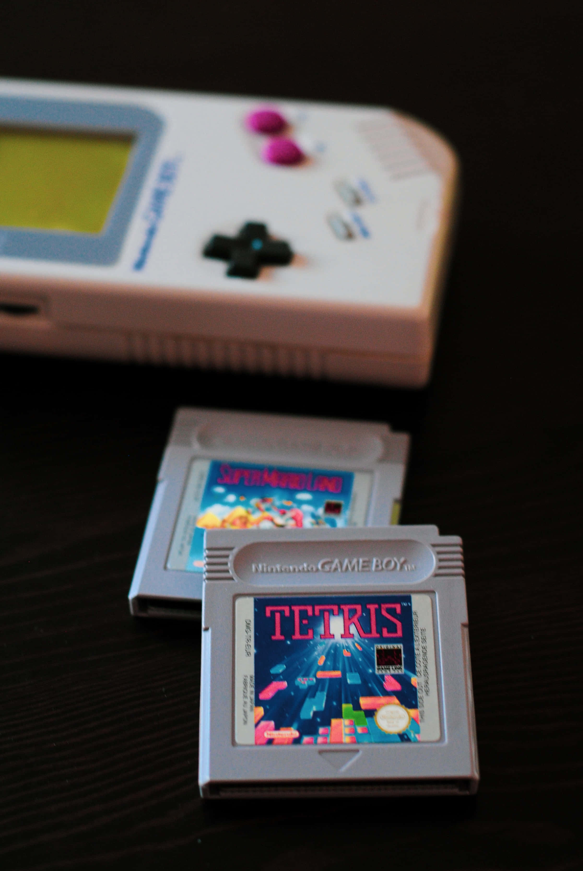 Gaming Consoles 桌布 壁紙 — 一部白色任天堂Game Boy，上面有兩張遊戲卡帶俄羅斯方塊和超級瑪利歐樂園，放在深色木質表面上。