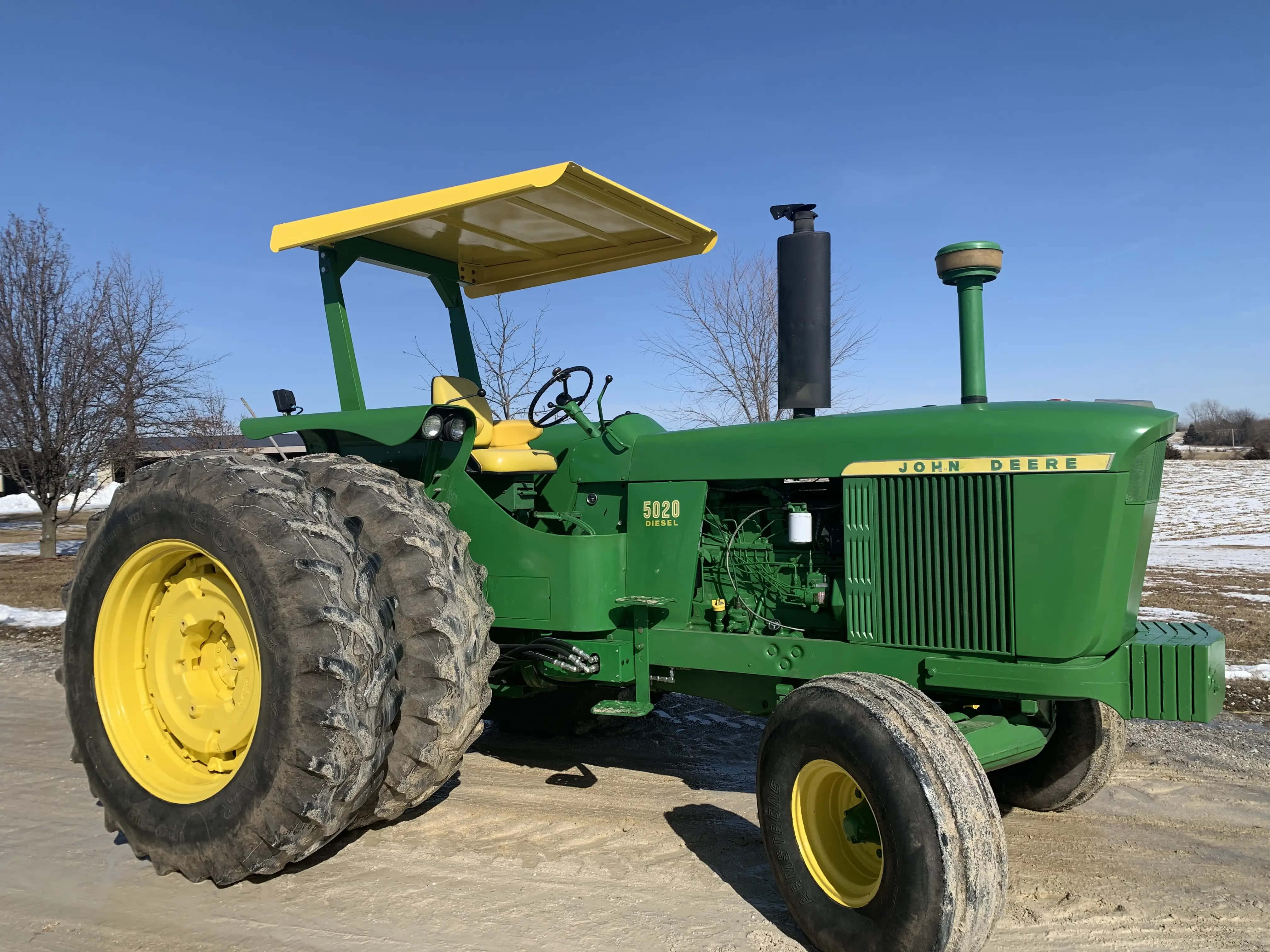 John Deere 桌布 壁紙 — 一部經典綠色和黃色的約翰迪爾5020拖拉機在晴朗藍天下自豪地矗立在白雪覆蓋的農場庭院中。