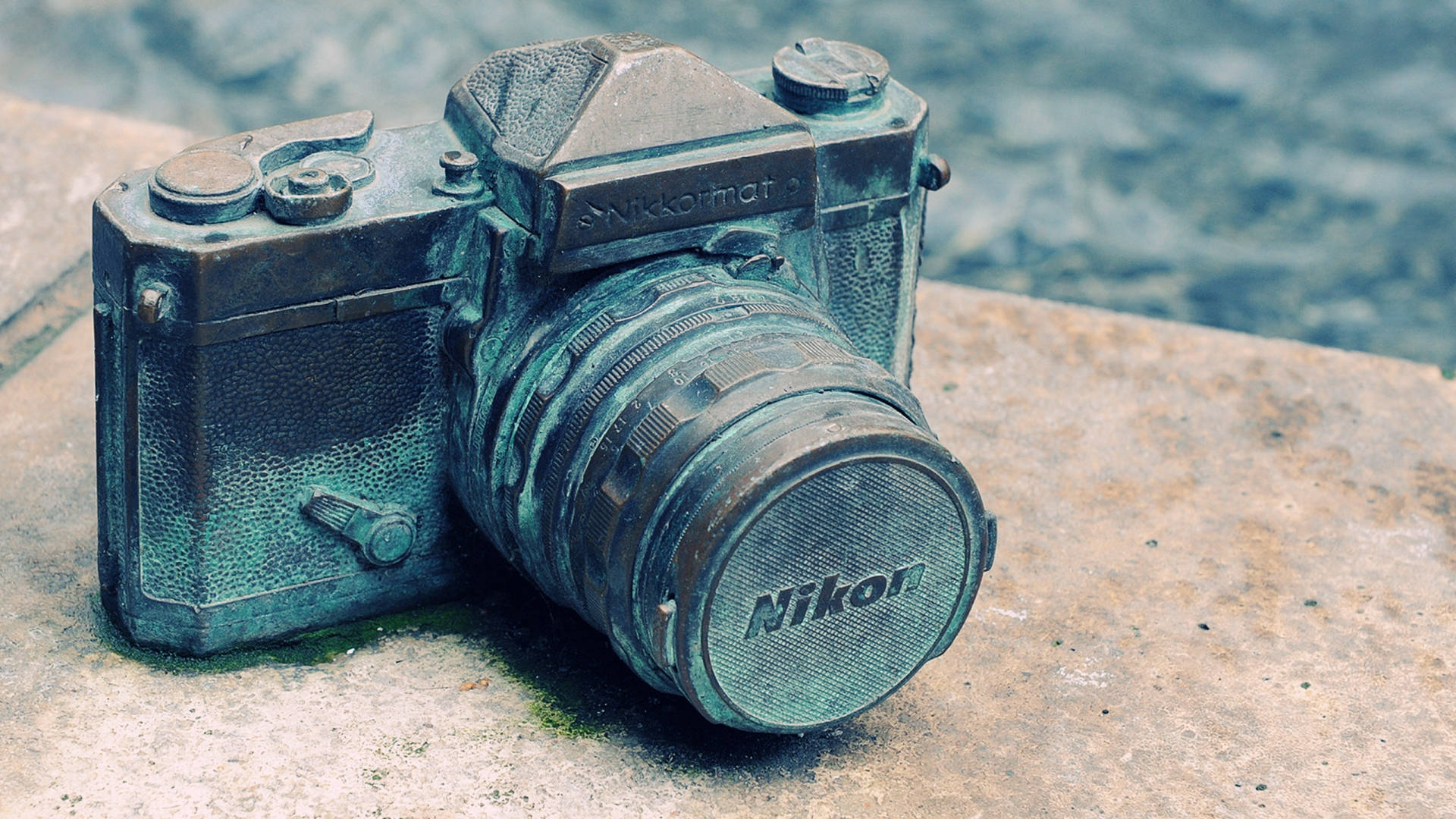  壁紙 — 一部經風化的復古Nikon相機擱在石架上，旁邊是漣漪的水面，機身覆蓋著青綠色的銅鏽。