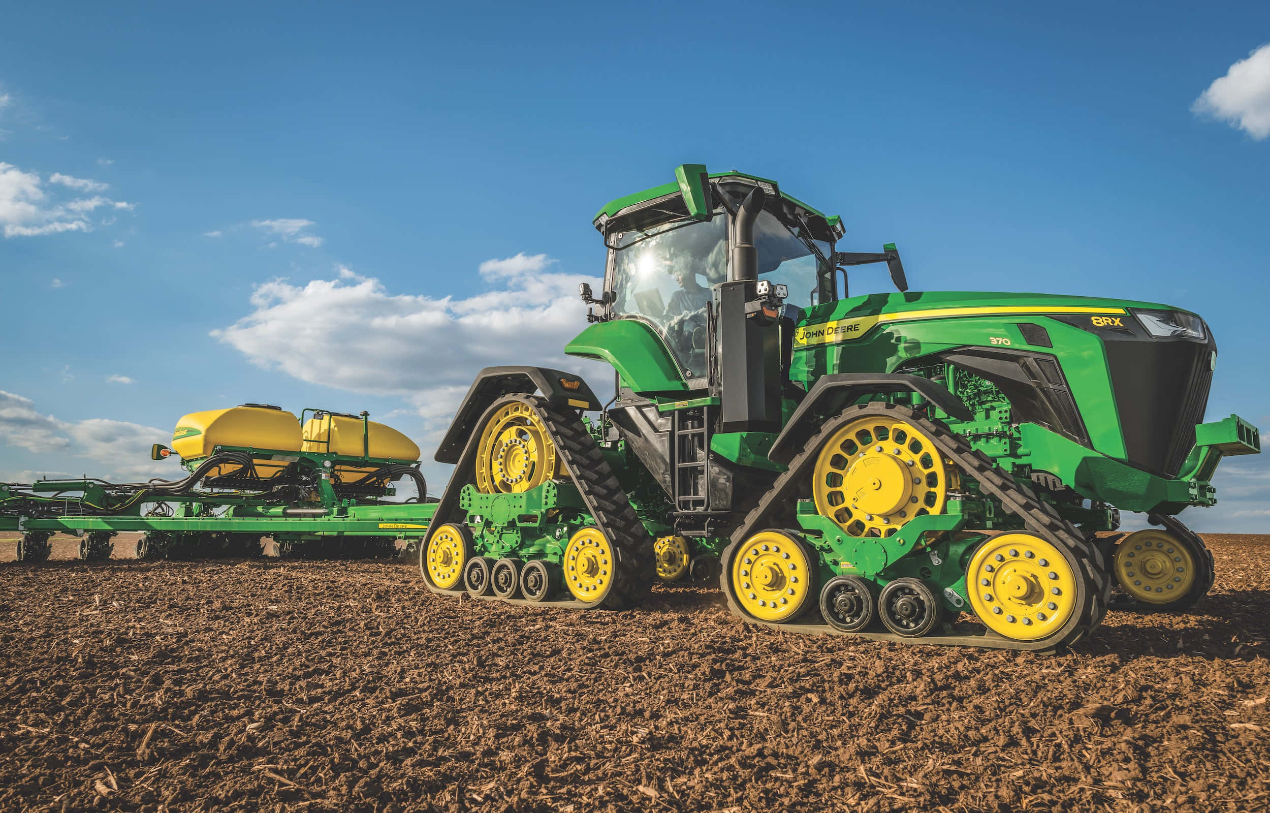 John Deere 桌布 壁紙 — 一部綠色和黃色的約翰迪爾履帶拖拉機在晴朗藍天和零星白雲下，拖著播種機穿過剛犁好的棕色田地。