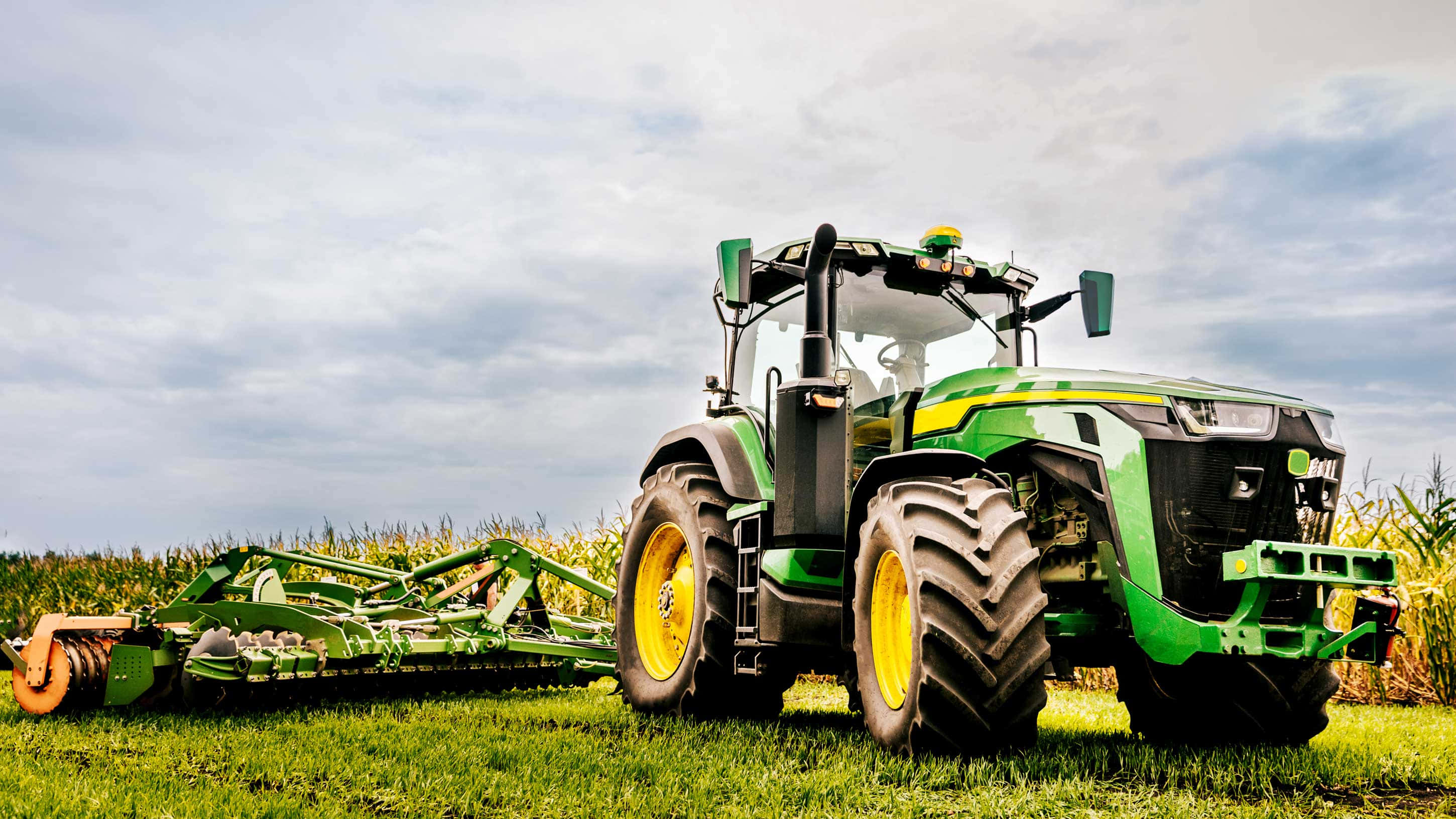 John Deere 桌布 壁紙 — 一部綠色和黃色的約翰迪爾拖拉機帶着一個大型犁，站在多雲天空下的草地上。